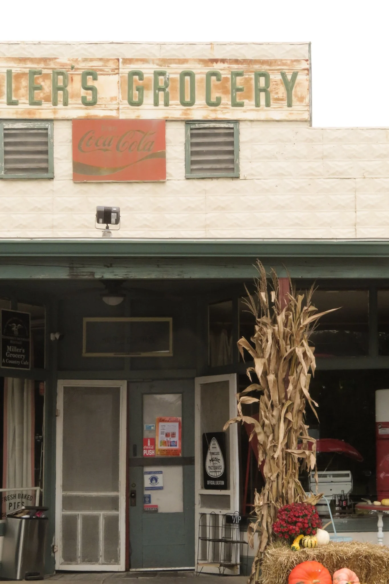 The exterior of a grocery store with a sign that reads "Grocery," and a Coca-Cola sign below it. In front of the store, there is a decorative display with autumn-themed items including a scarecrow-like figure made of dried corn stalks, pumpkins, and 