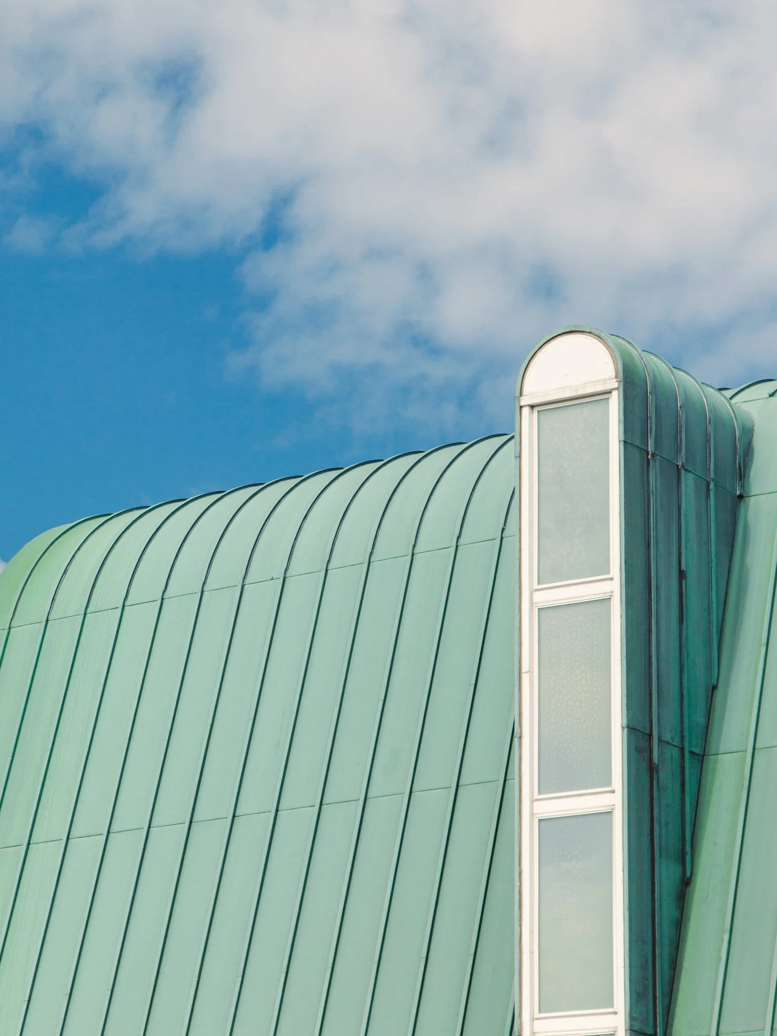 Close-up of a building with a green metal roof and a tall, narrow window with a curved top, set against a blue sky with scattered clouds.