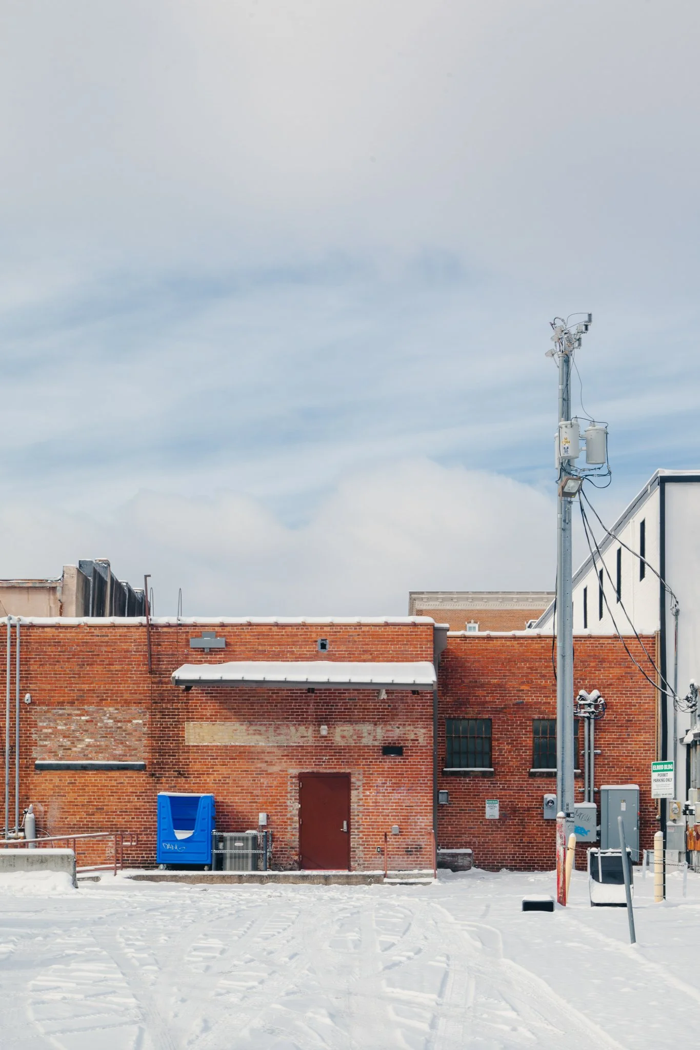 Snow-covered urban alley with red brick building, utility pole, and scattered equipment.