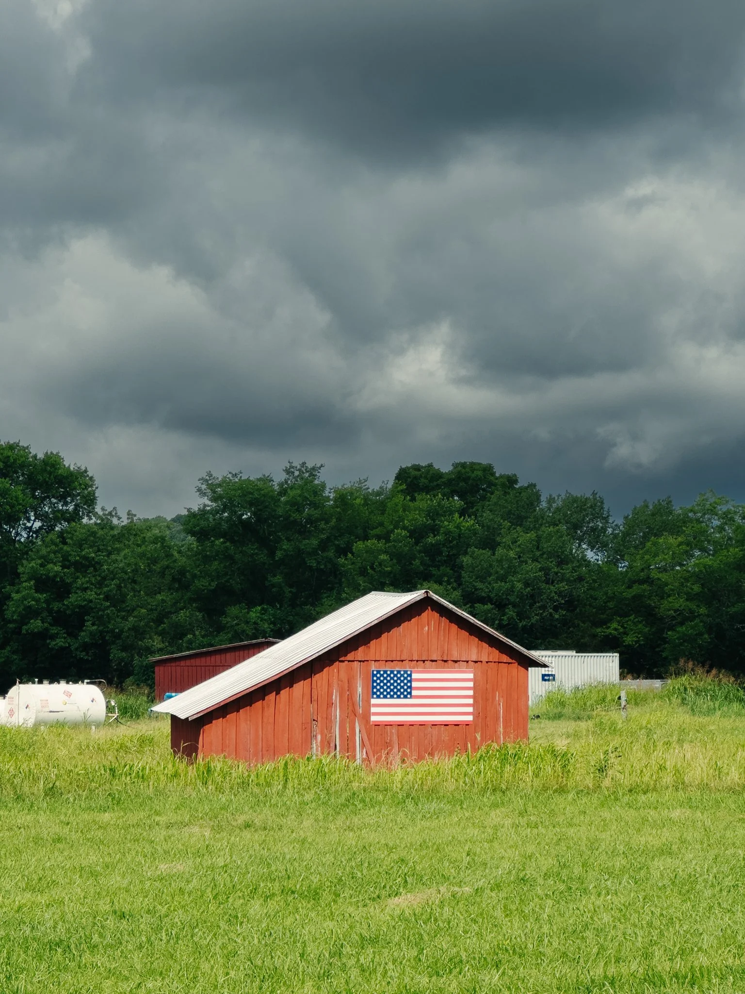 A red barn with an American flag on its side, surrounded by green grass, with a forested hillside and dark cloudy sky in the background.