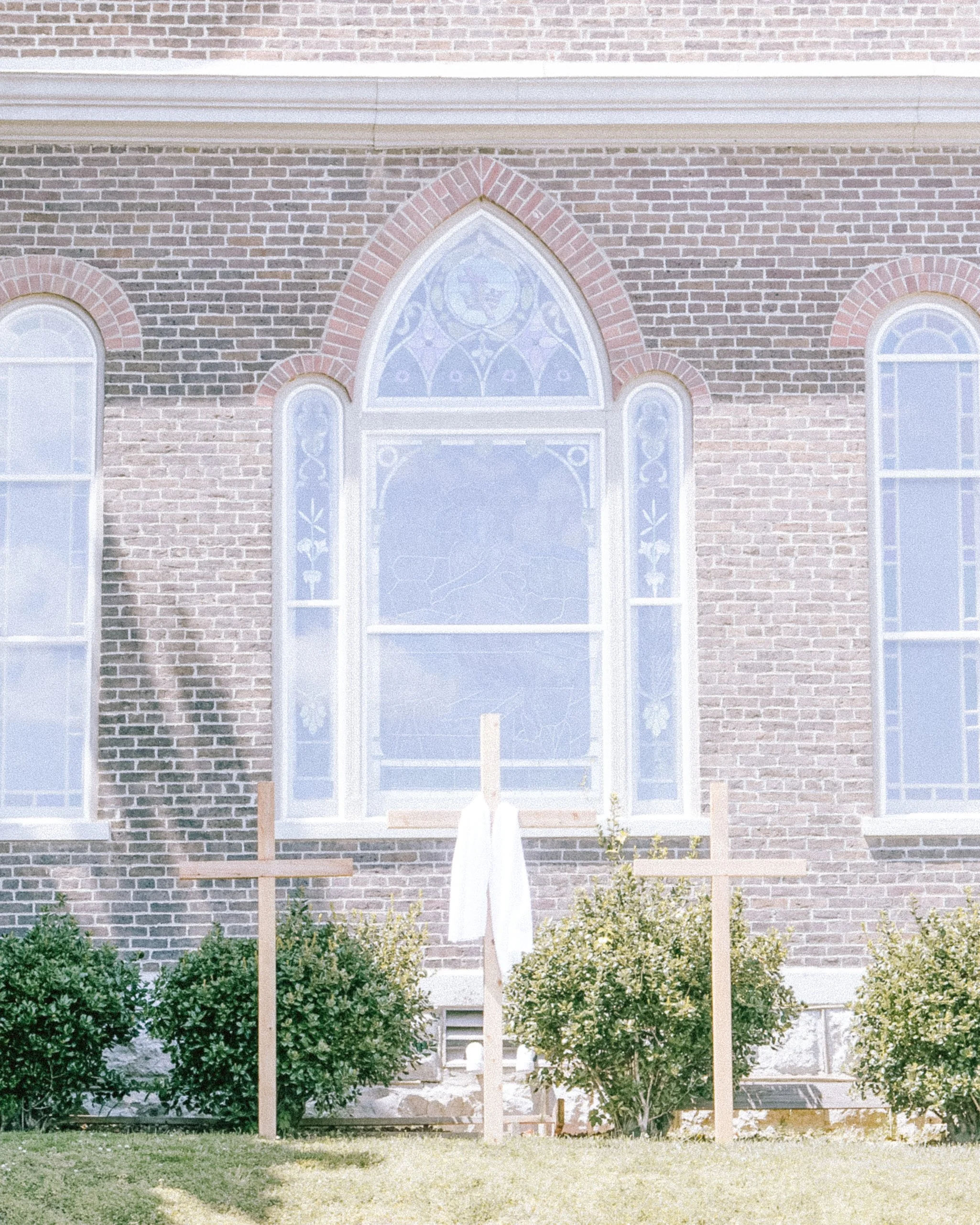 A brick church building with arched stained glass windows and a wooden cross, trees and bushes in front, and a white cloth draped over a wooden stand in the front yard.