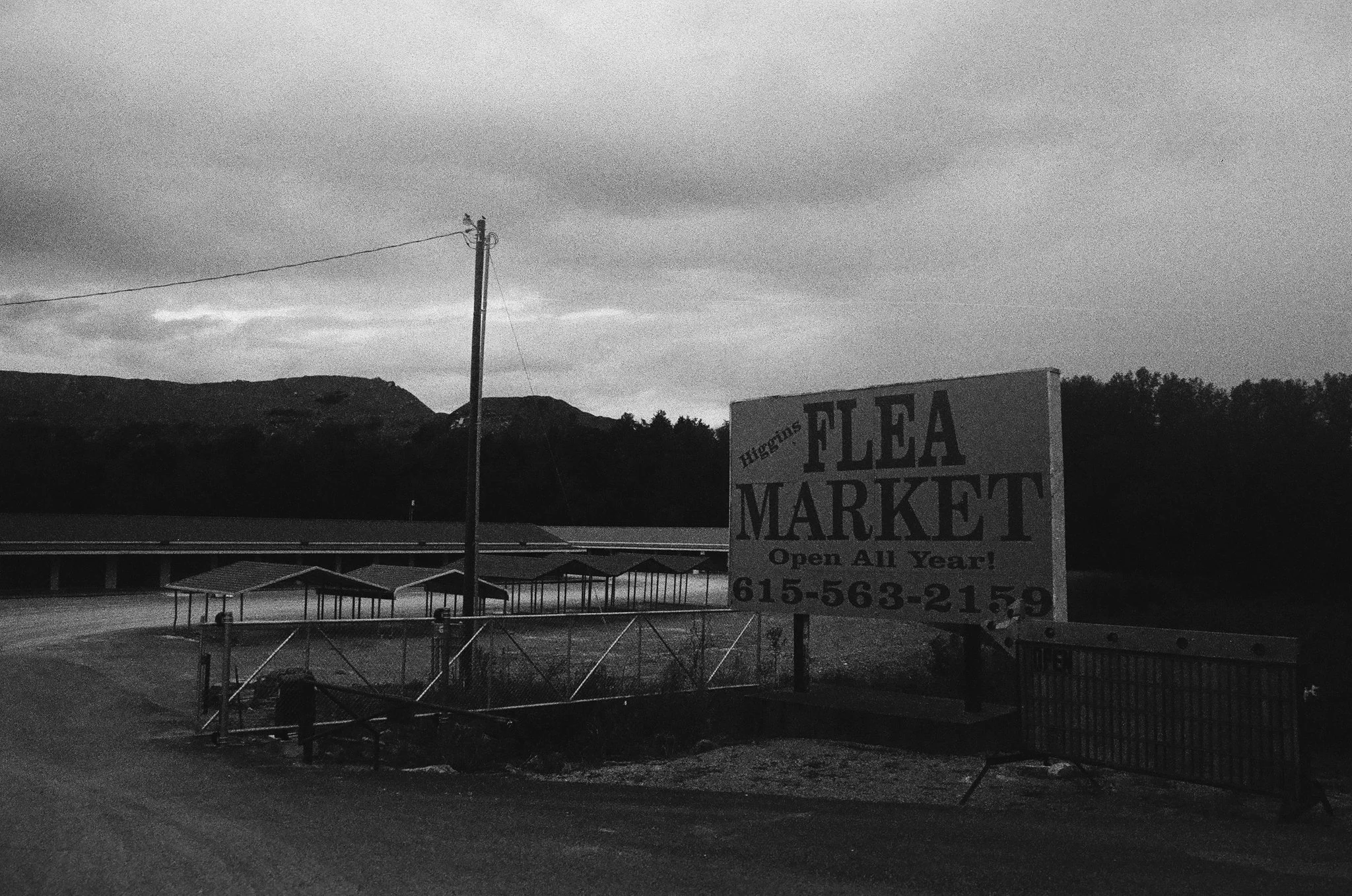 A parking lot with a large sign for Flea Market, open all year, with a phone number, surrounded by mountains and trees, under an overcast sky.