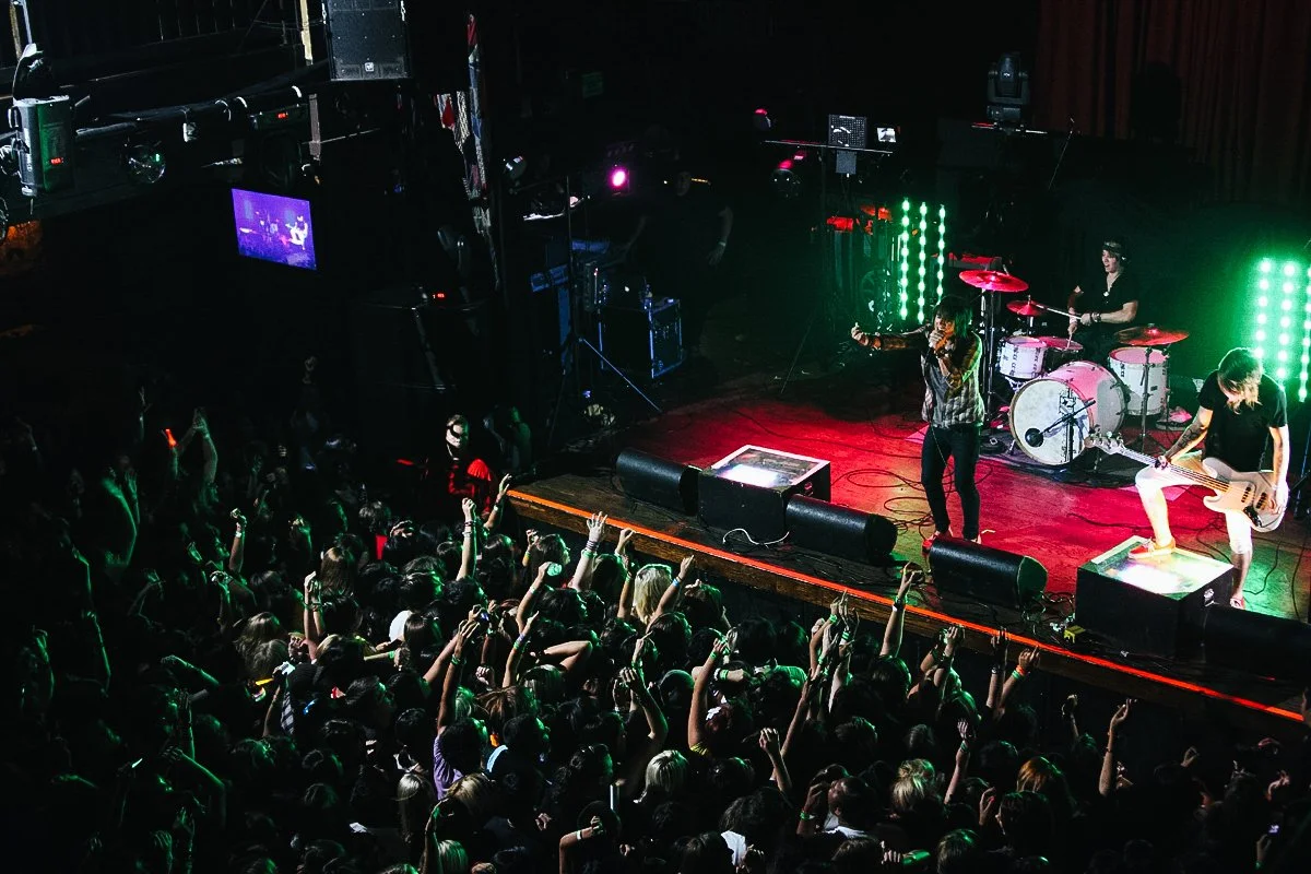 A band performing on stage with a singer, drummer, and guitarist in front of an audience at a concert venue, with colorful stage lighting.