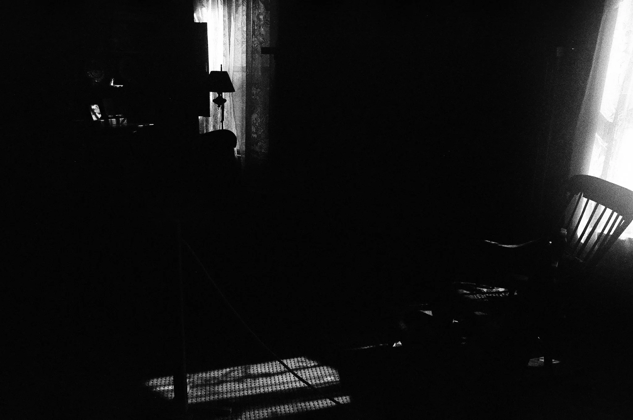 A dark room with light coming through window blinds, casting patterned shadows on the floor and furniture including a chair, lamp, and side table.