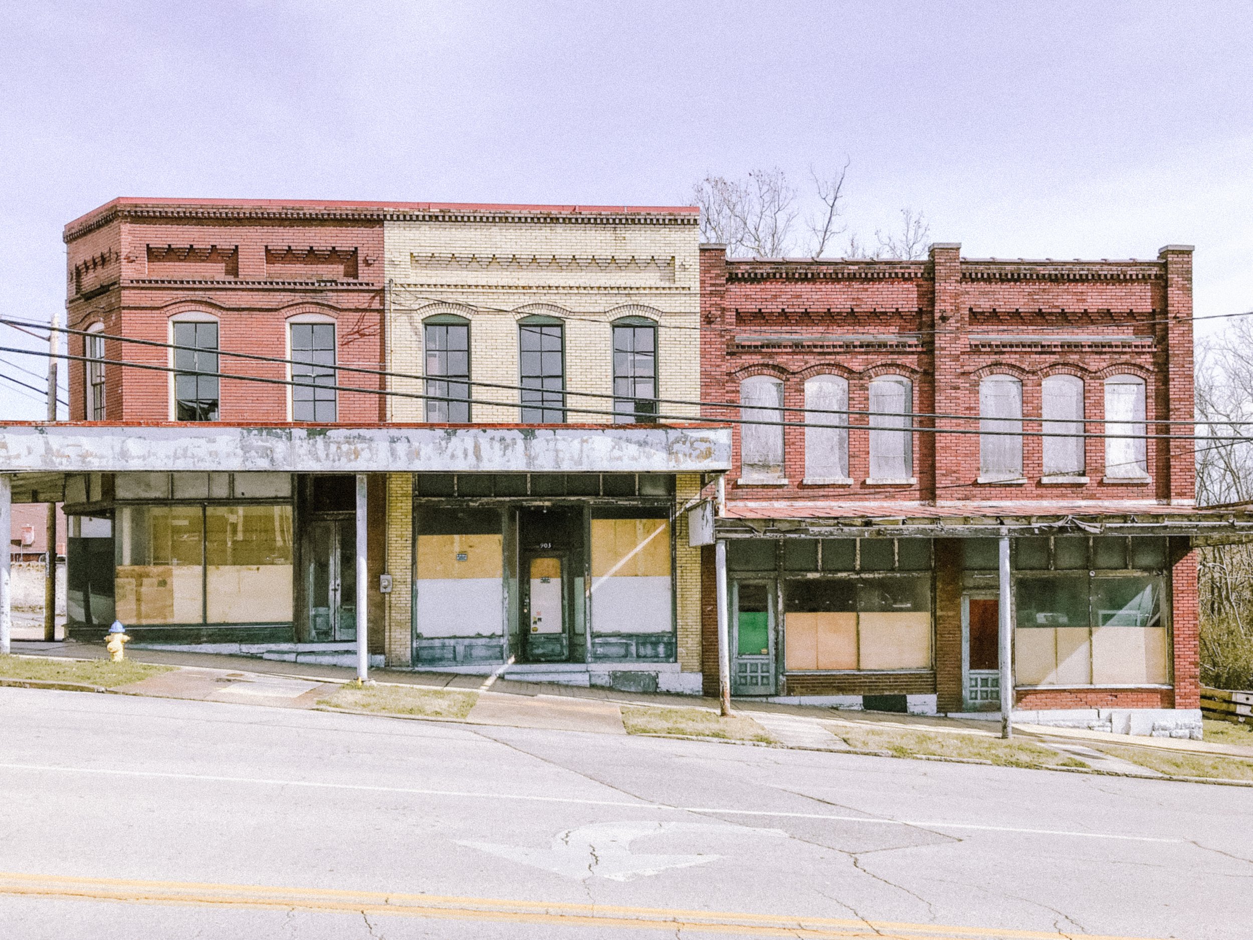 An old, abandoned building with brick walls and boarded-up windows, located on a city street with power lines and a fire hydrant in the foreground.