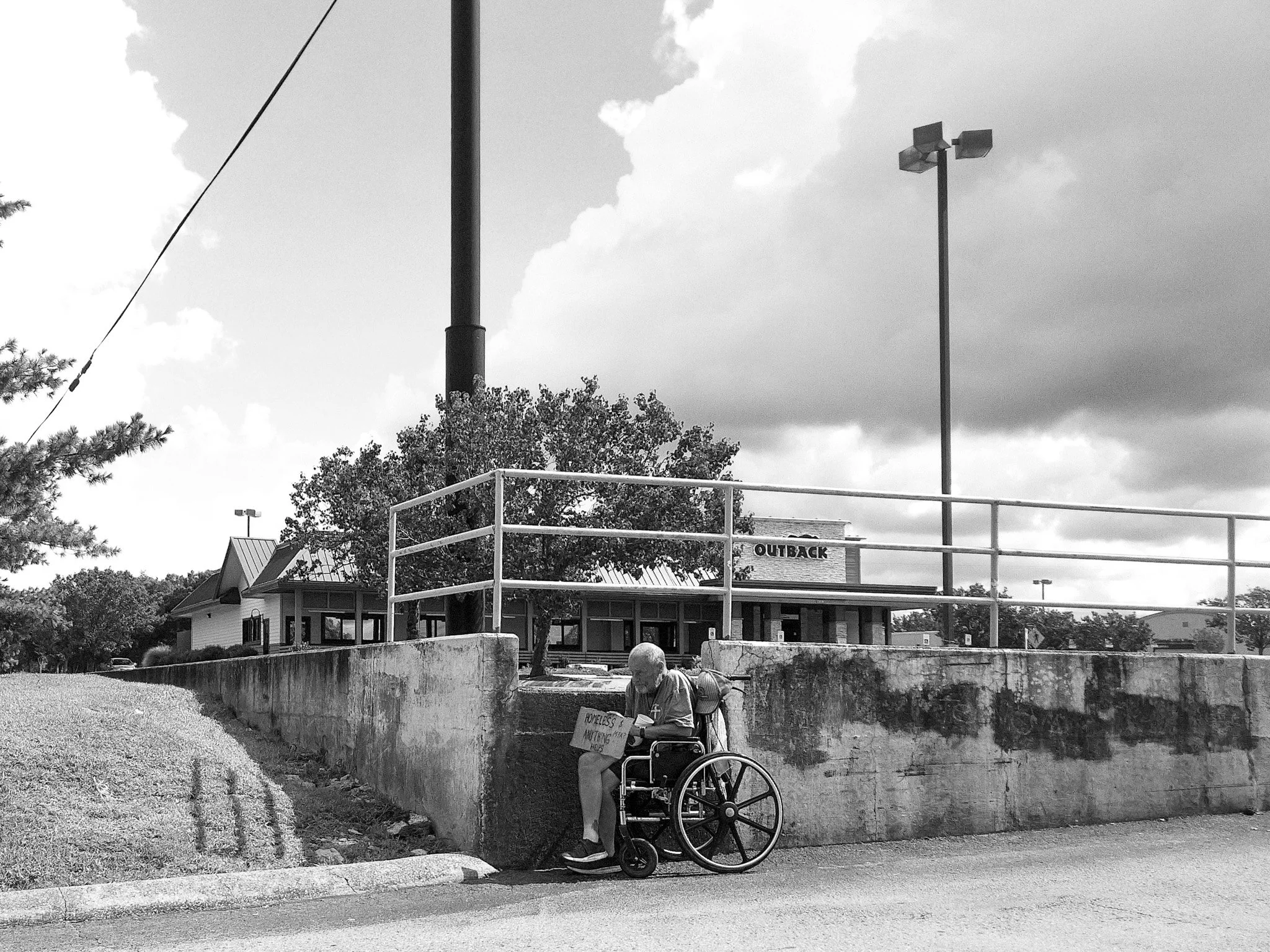 A person sitting in a wheelchair under a bridge, holding a cardboard sign that reads 'HOMELESS & NOTHING TO LOSE.' In the background, there is a building with the sign 'OUTBACK,' trees, and a light pole, with a cloudy sky overhead.