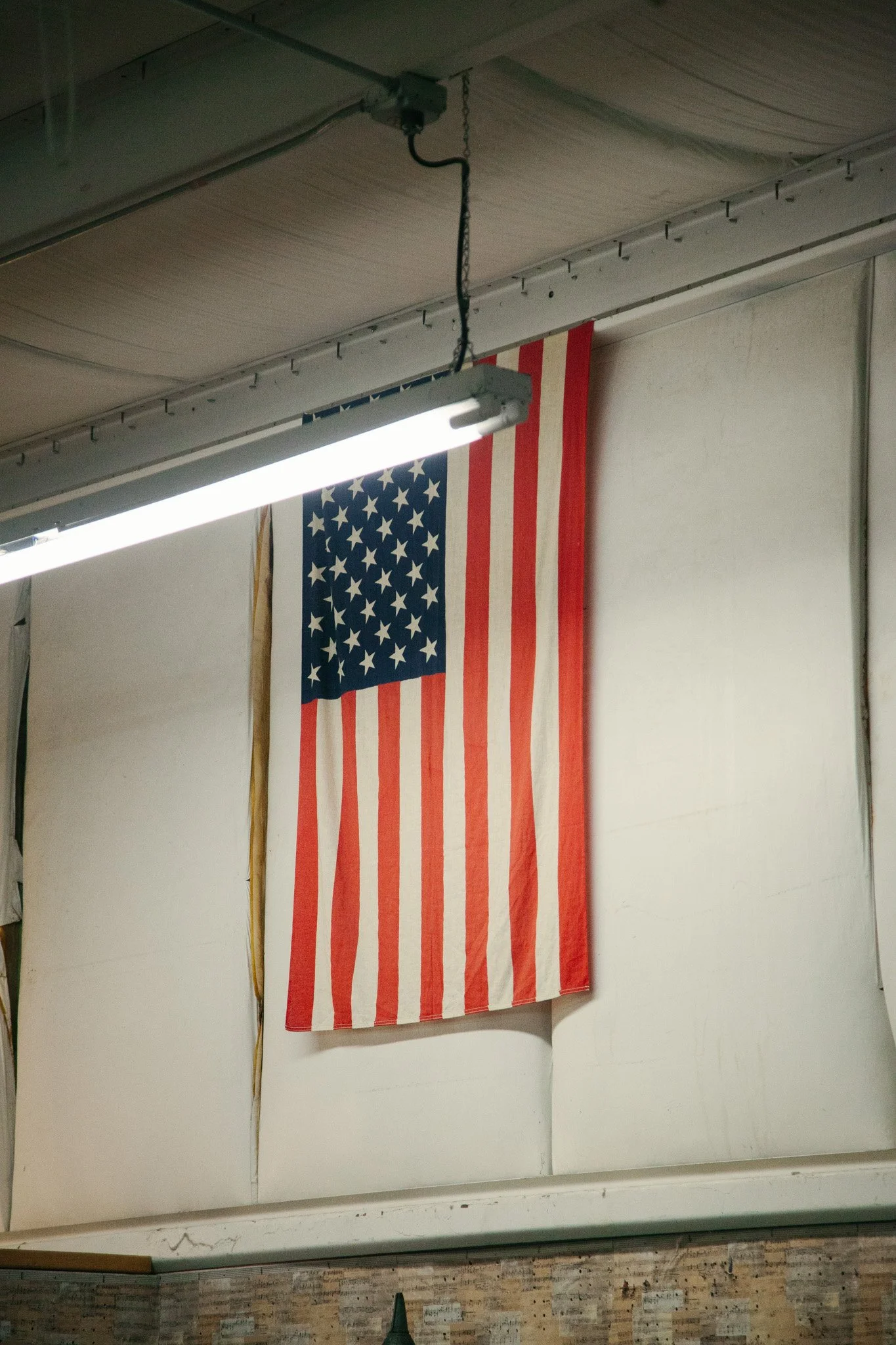 American flag hanging on a wall in an indoor setting, partially illuminated by a ceiling light.