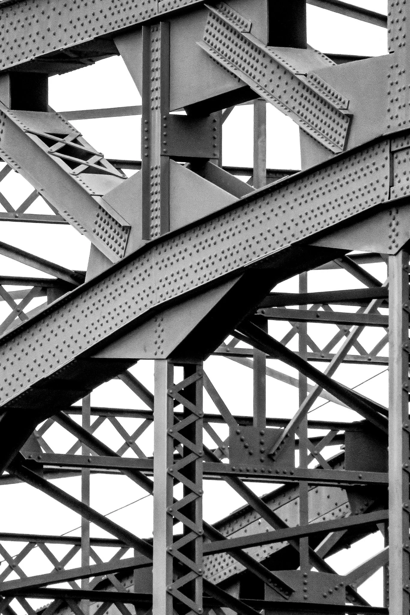Close-up of a section of an iron bridge showcasing intersecting beams and rivets, with structural support beams visible in the background, in black and white.