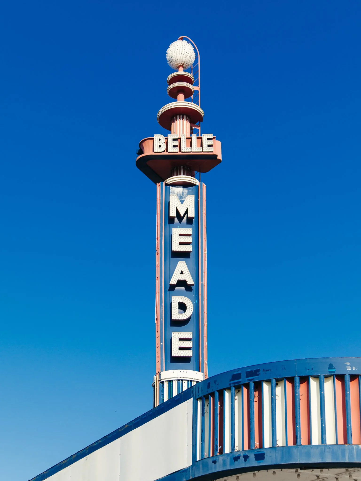 Tall neon sign with the word 'Belle' at the top and 'Mega' below, part of a vintage-style building with colorful vertical stripes and a curved balcony against a clear blue sky.