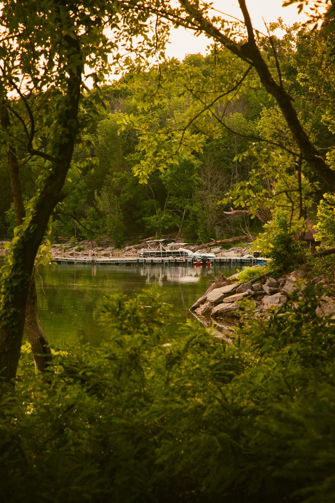 A peaceful river scene at sunset, surrounded by green trees and rocks, with a dock and boats moored along the shoreline.