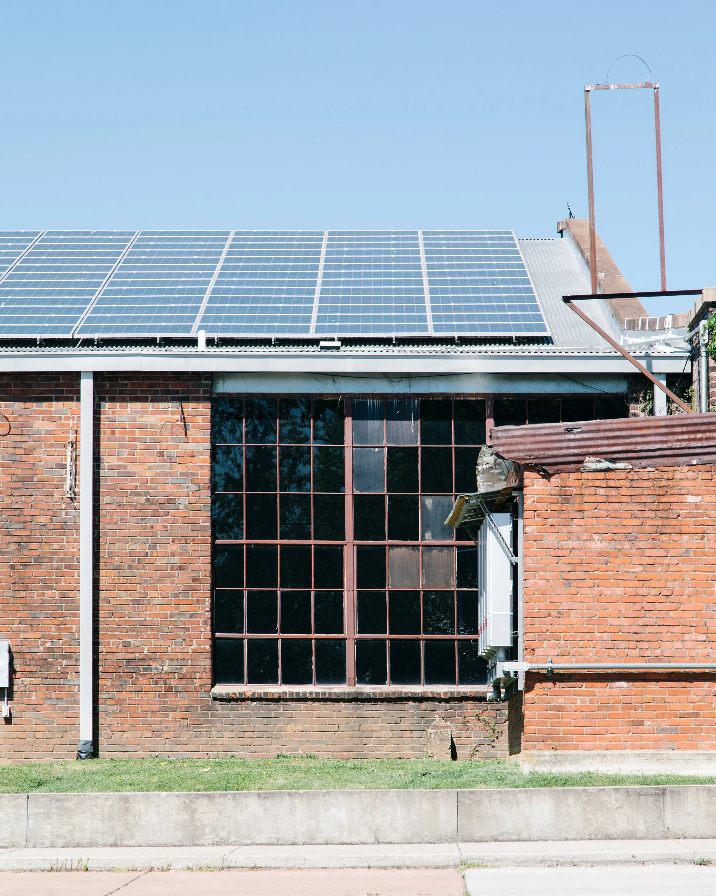 Building with solar panels on roof, brick exterior, large window, electrical equipment and storage tank on side, surrounded by a lawn and sidewalk.