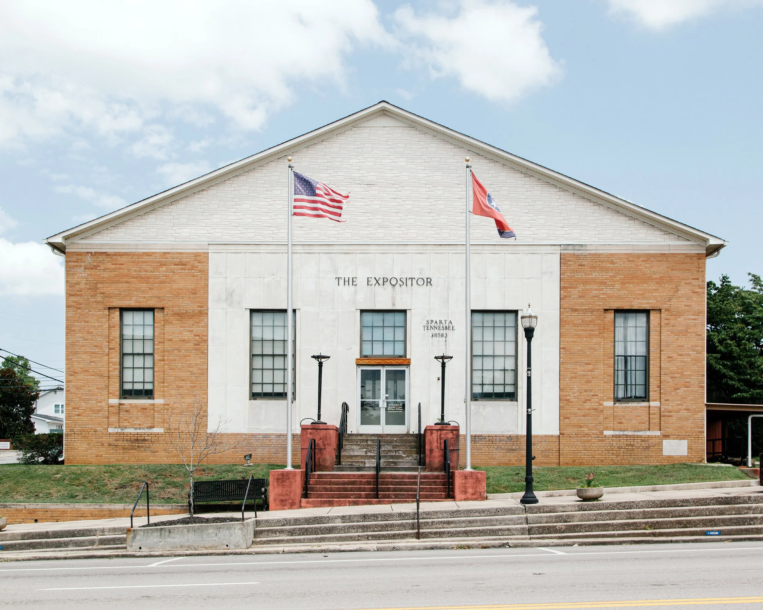 Front view of a building named 'The Expositor' in Sparta, Tennessee, with two flagpoles displaying the American and Tennessee flags, concrete stairs leading up to the entrance, and a black park bench in front.