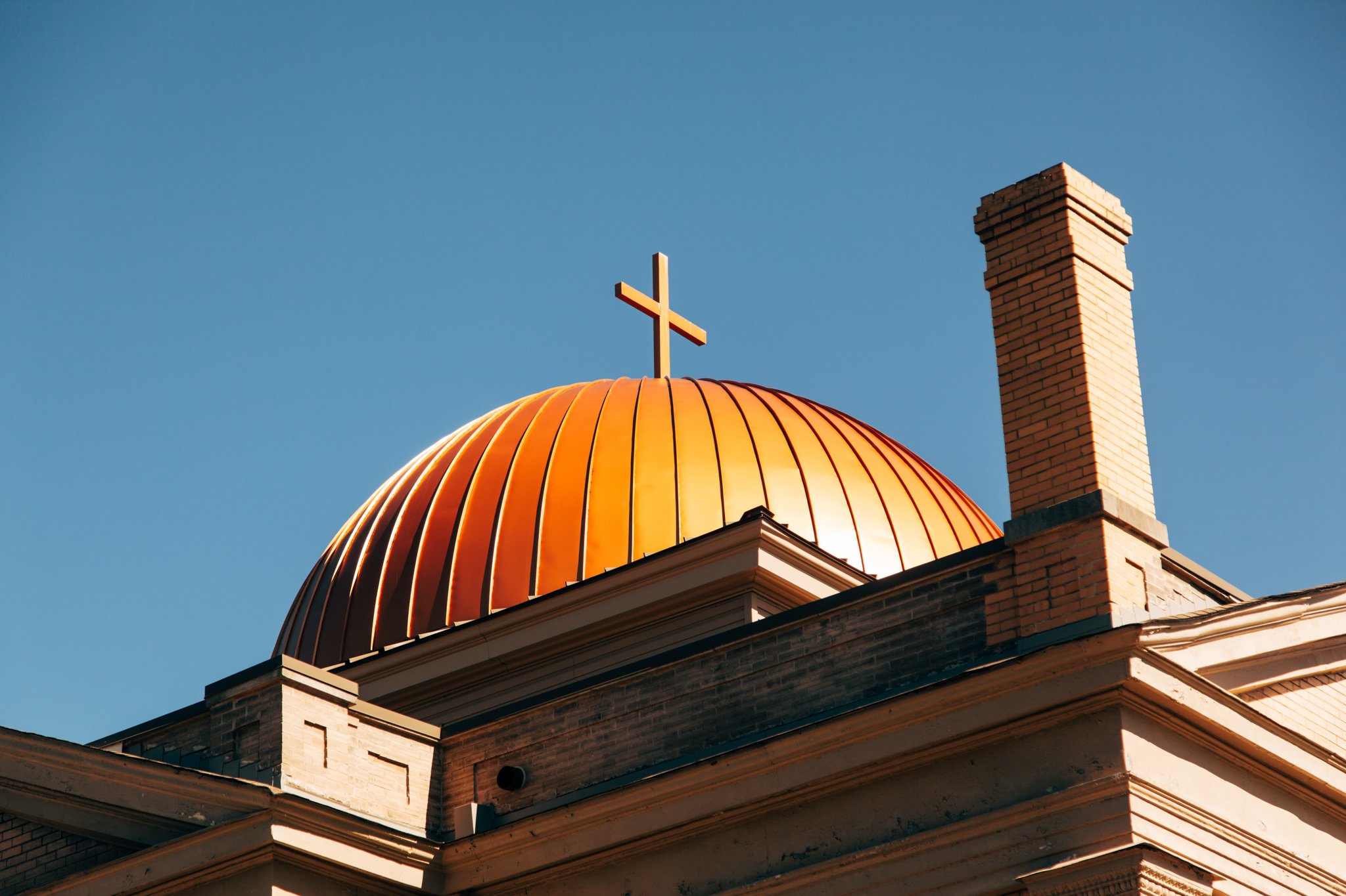 Close-up of a church building's golden domed roof with a cross on top, brick chimney, and architectural details against a clear blue sky.