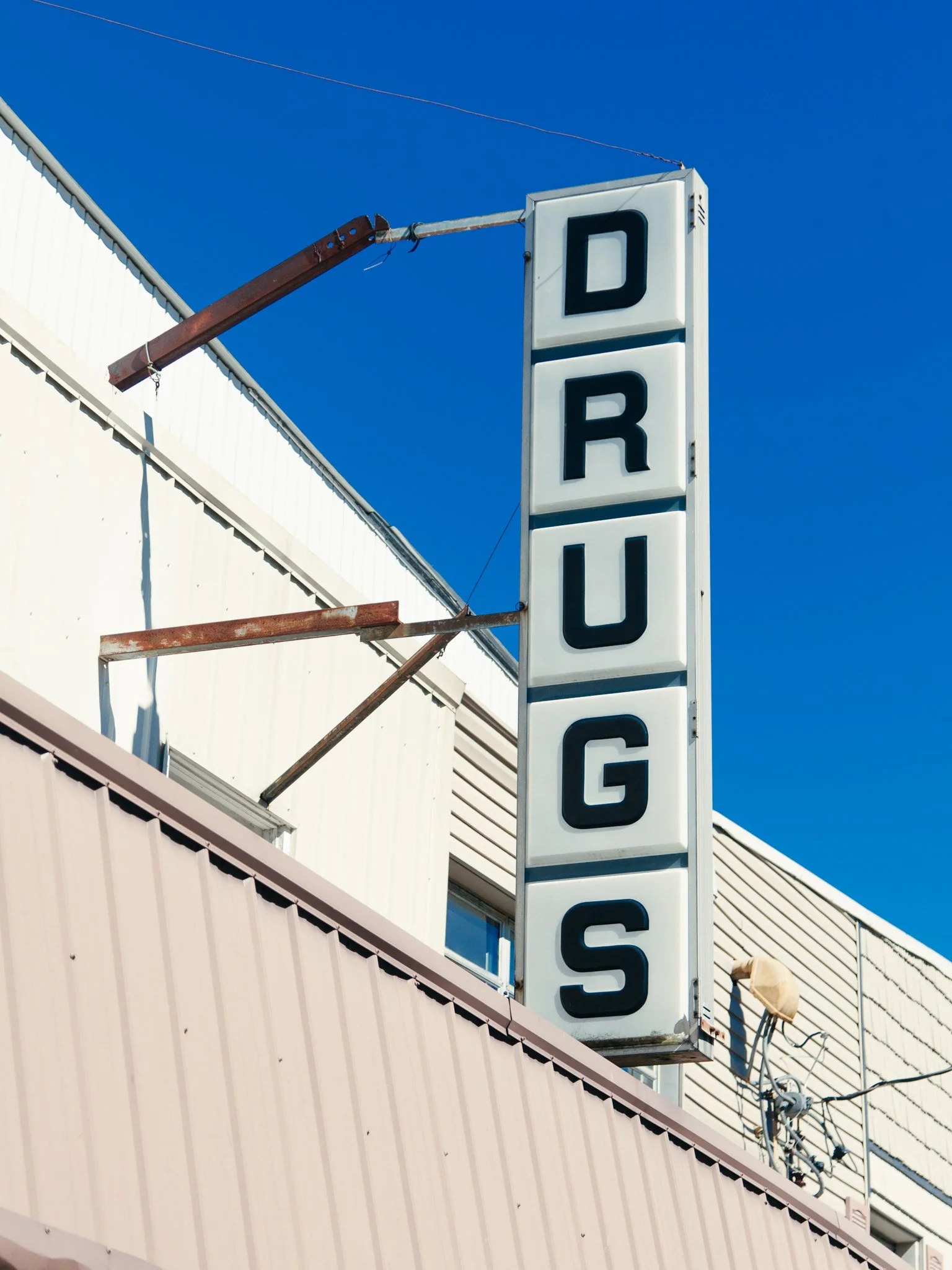 A white vertical sign with black letters spelling 'DRUGS' hangs from a rusty metal bracket against a bright blue sky, attached to a beige building with metal siding.