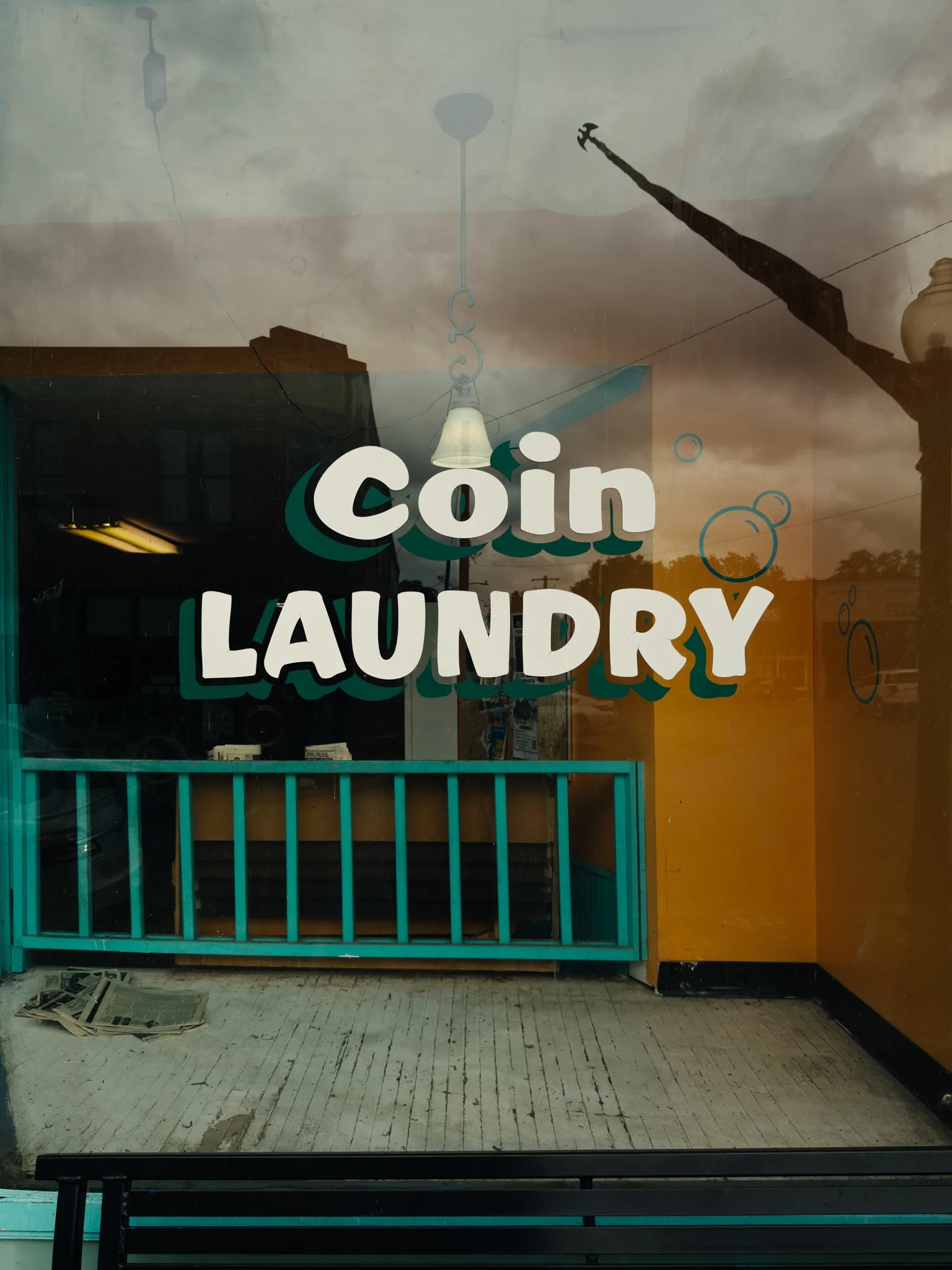 View of the inside of a laundry with a sign on the window reading 'coin laundry'. The laundry appears empty with a cash register visible inside and newspapers on the floor. The floor is whitewashed wood with some wear.