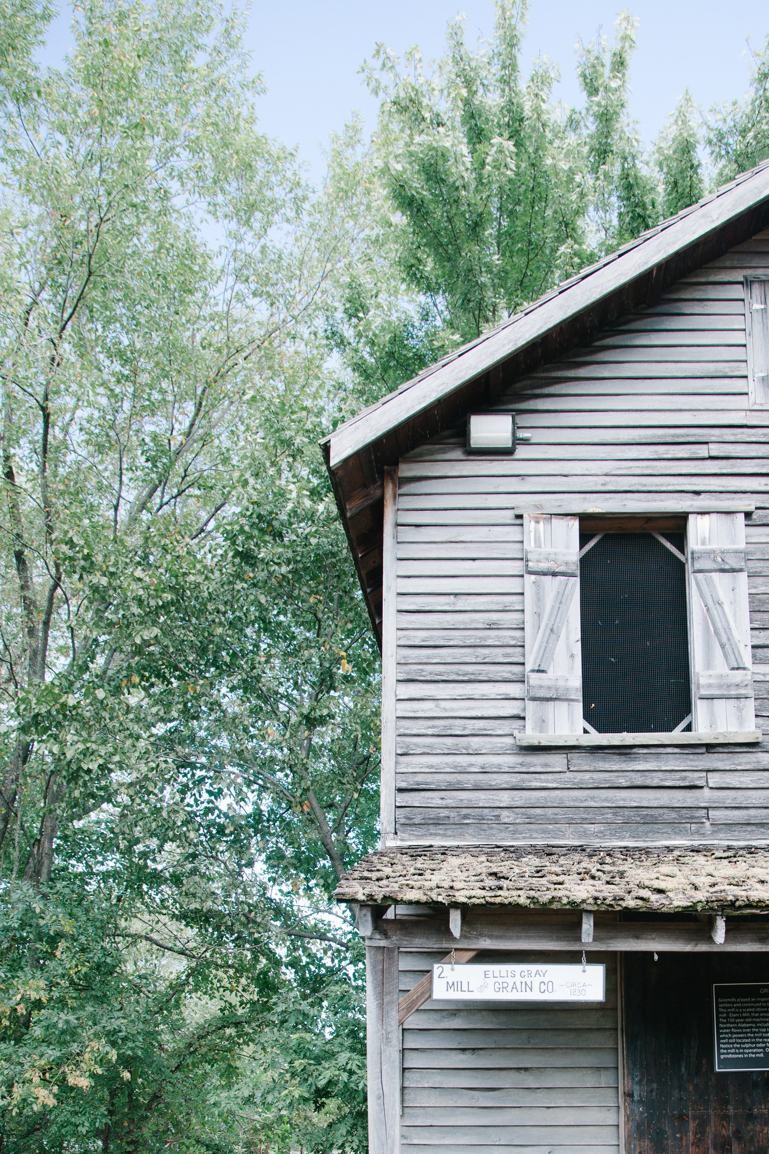 An old wooden mill building with a shingles roof and shuttered window, partially visible. Trees with green foliage are behind the building. A white sign hangs on the front, reading 'Mill Ellis Gray Grain Co. circa 1830'.