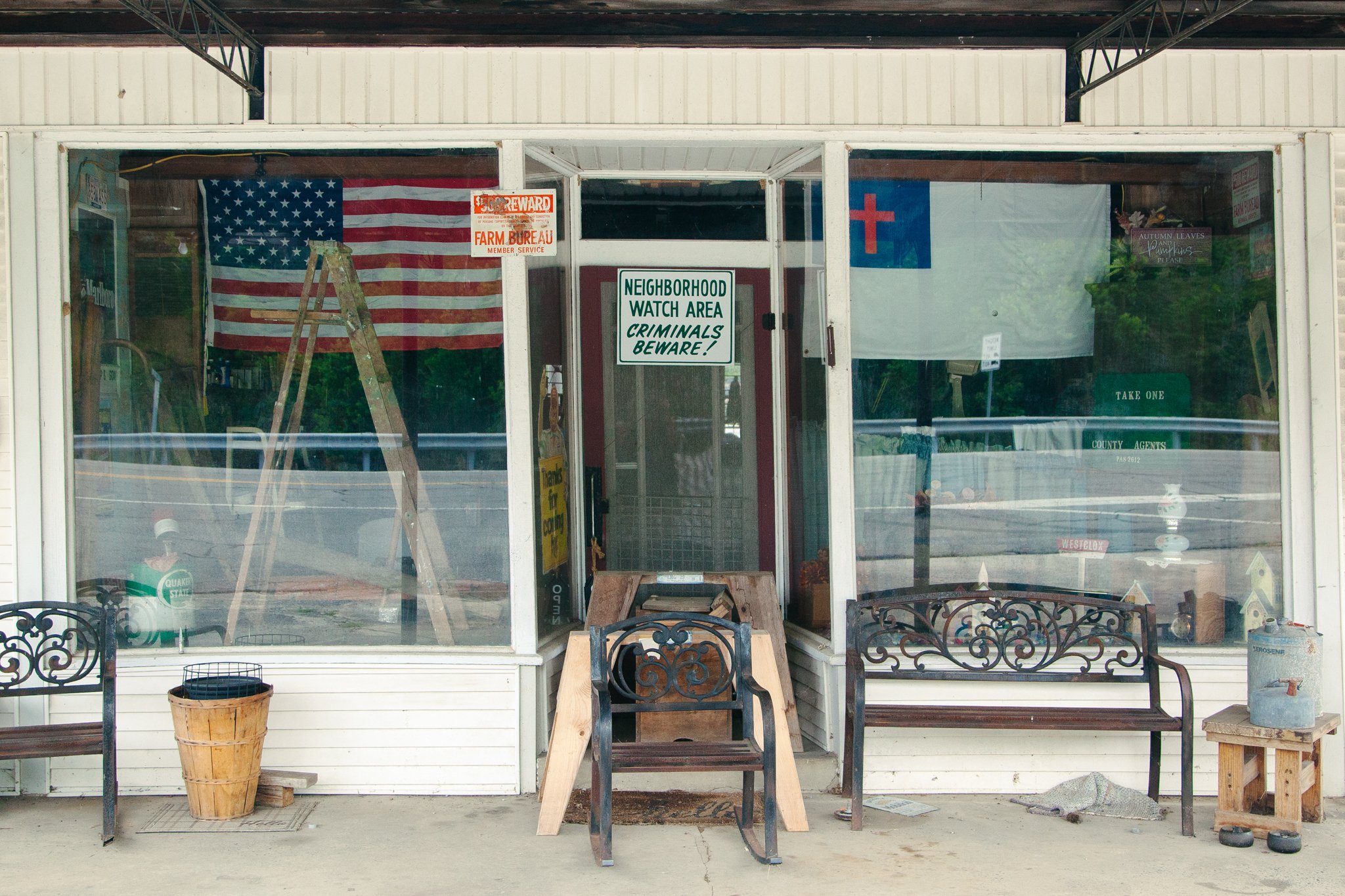 View through a storefront window showing American flag, a Christian cross flag, and signs including one that says 'NEIGHBORHOOD WATCH AREA CRIMINALS BEWARE!'. Inside the store, there are ladder, books, a can of propane, and decorative items.