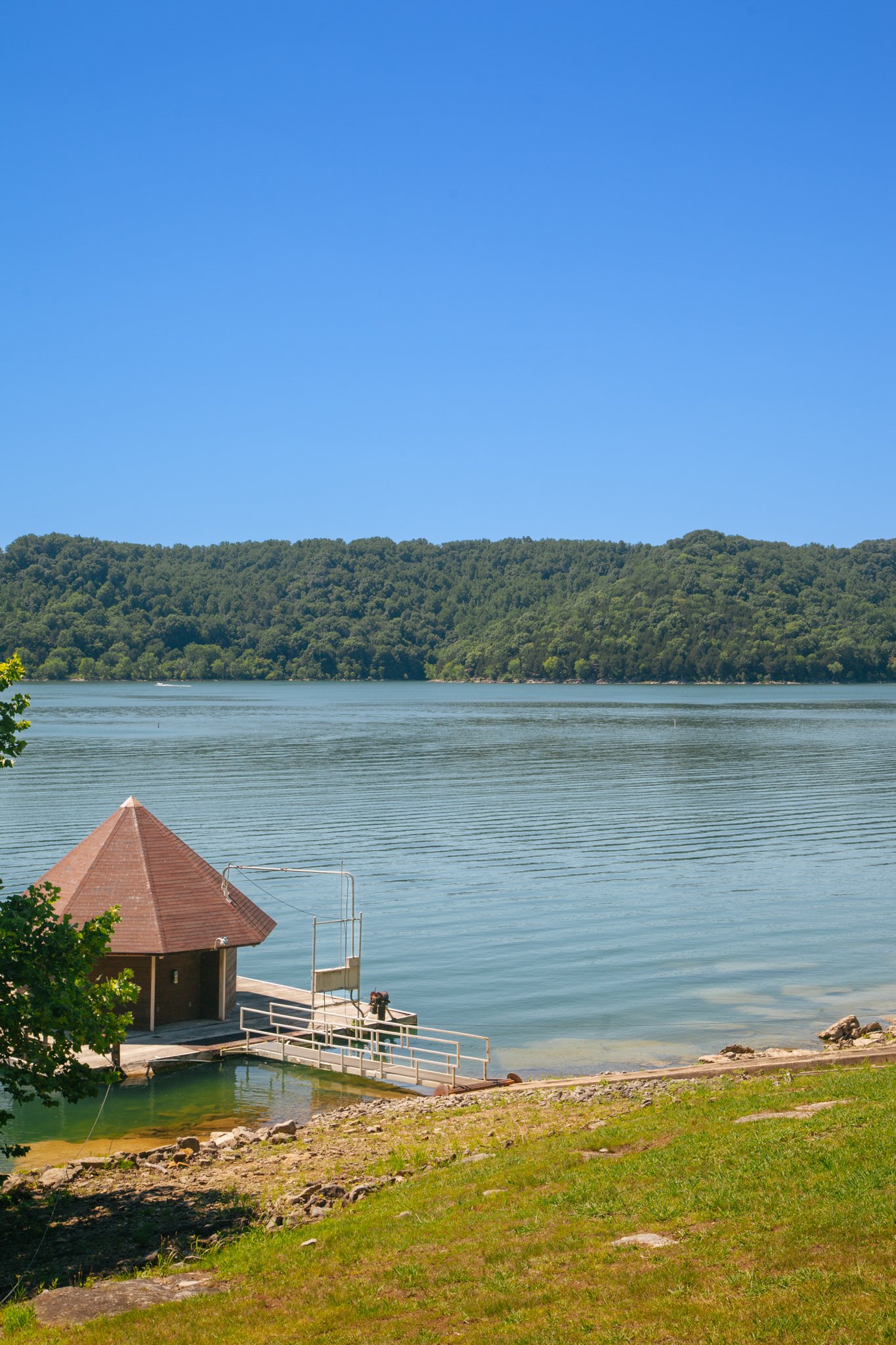 A lake surrounded by green trees and hills under a bright blue sky. A small brown building with a cone-shaped roof and a deck with a railing is in the foreground near the water.