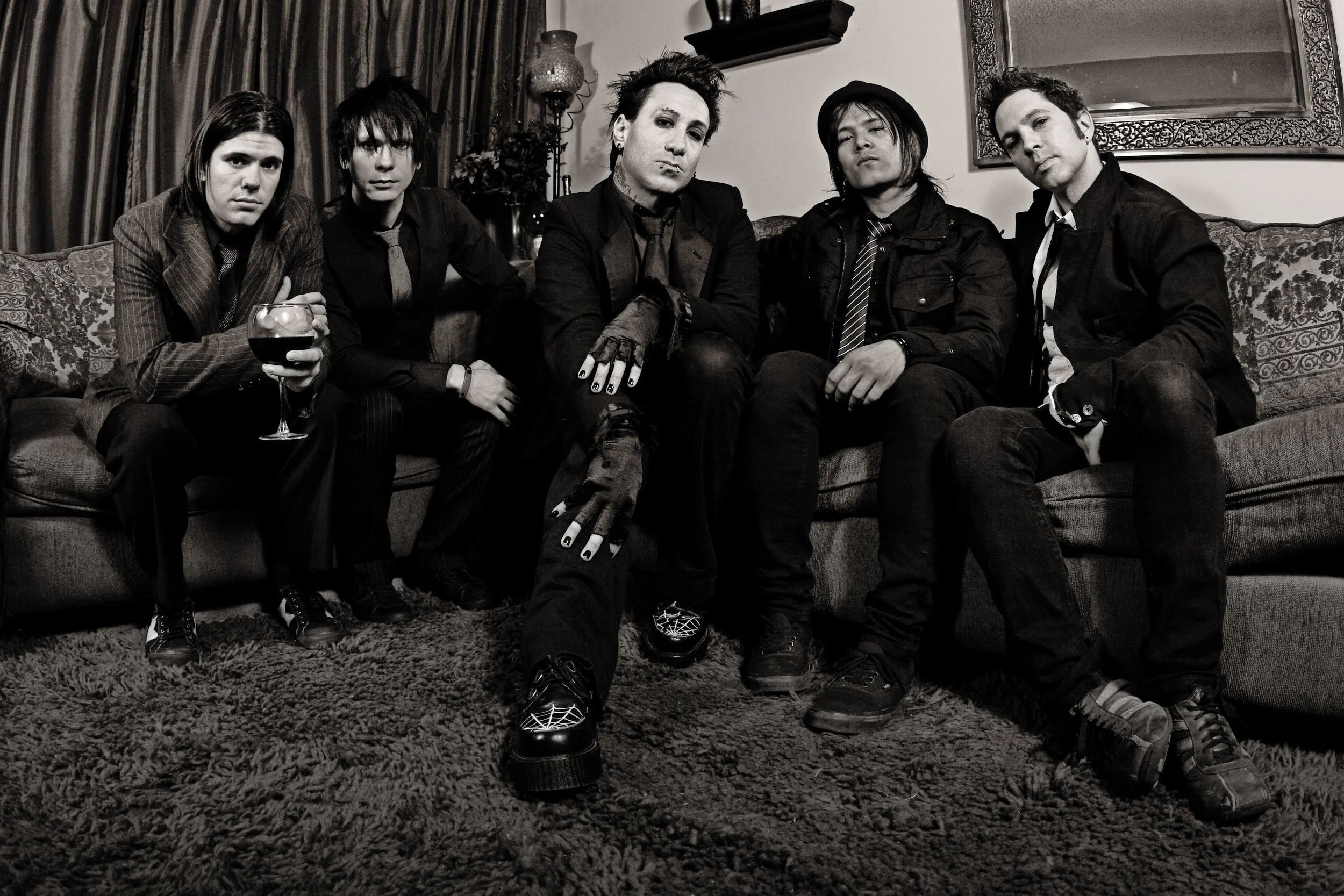 A group of five young adults sitting on a vintage-style sofa in a dimly lit room, dressed in alternative fashion, with serious expressions, one holding a glass of red wine, and the background featuring ornate curtains, a decorative lamp, and wall art