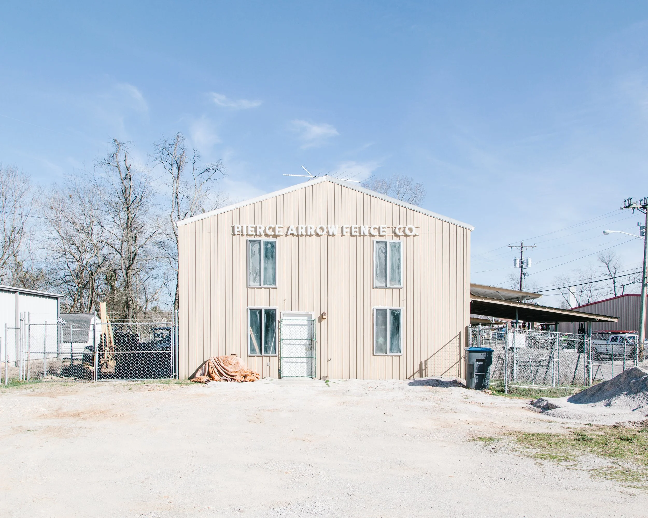 A beige building with a sign that reads 'Pierce Arrow Fence Co'. It has multiple narrow windows and is surrounded by a chain-link fence. In front of the building, there is a dirt ground with some debris and a black trash bin. Behind, there are leafle