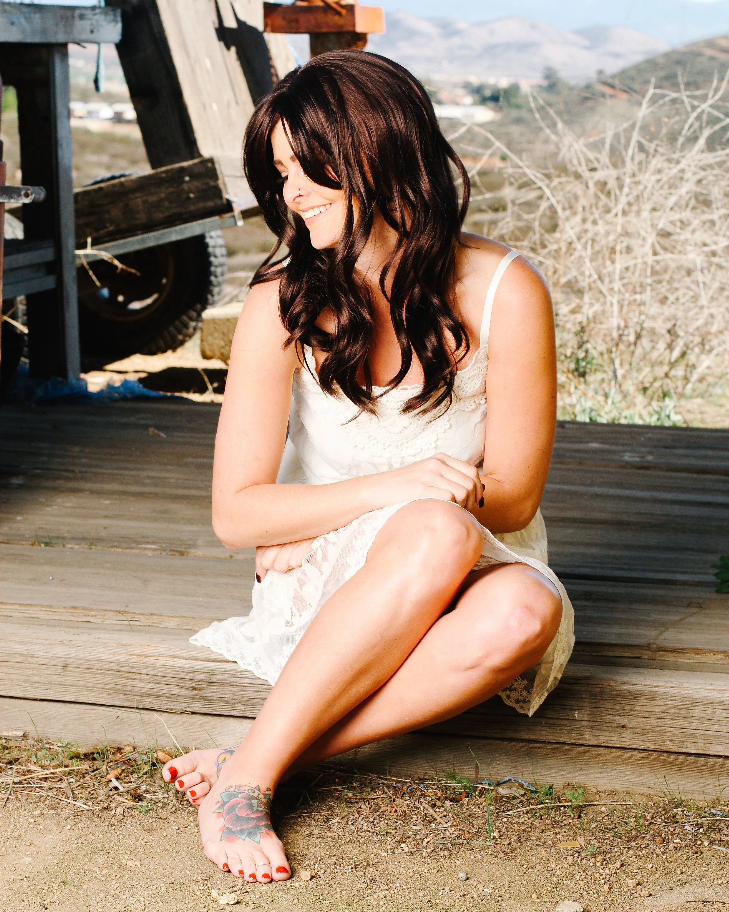 A woman with long dark hair, wearing a white lace dress, sitting outdoors on a wooden surface, smiling and looking down. She has a colorful tattoo on her foot and red painted toenails. The background shows a landscape with hills and dry bushes.
