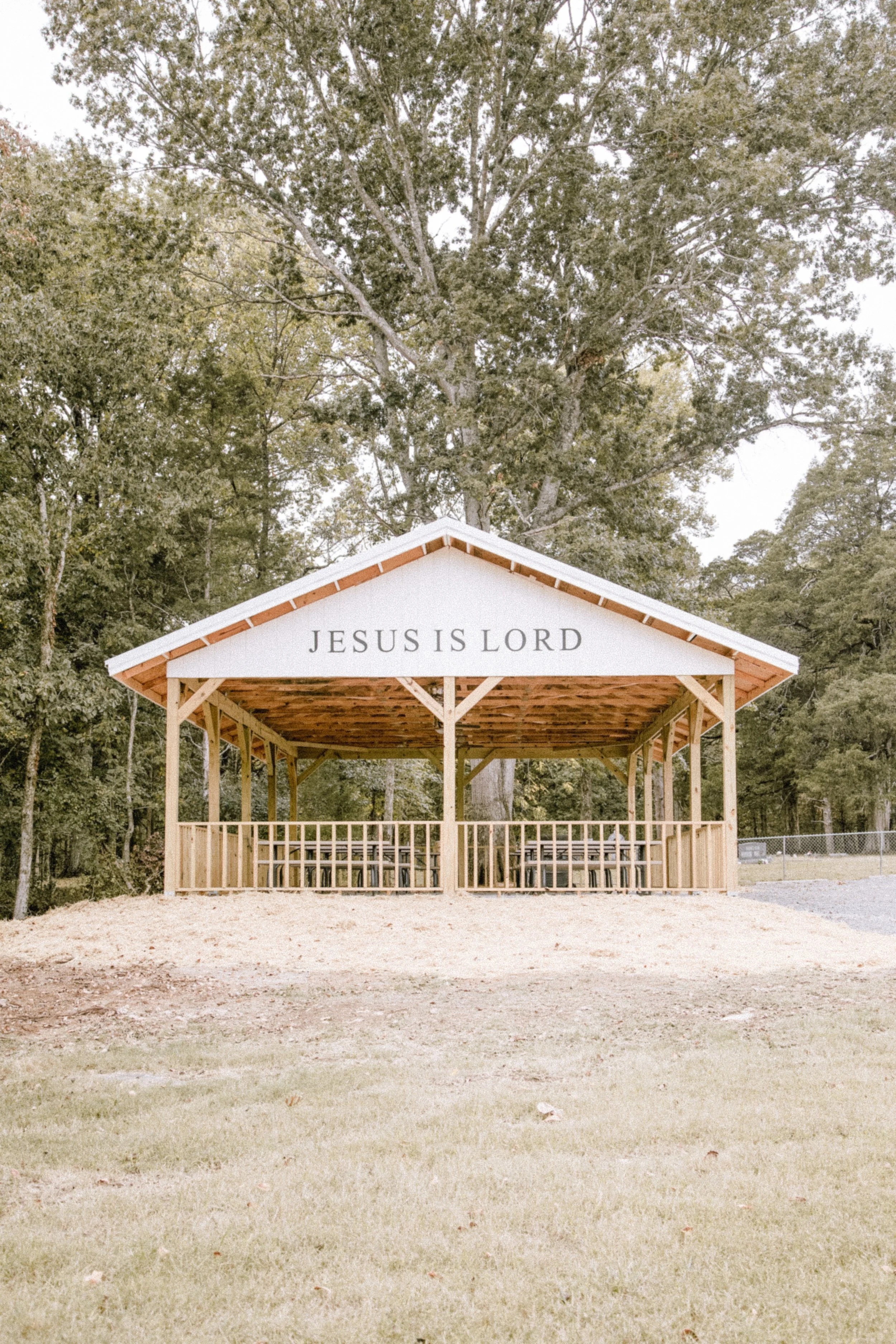 A small outdoor pavilion with a wooden structure and white roof, displaying the words 'JESUS IS LORD', situated in a grassy area with trees in the background.