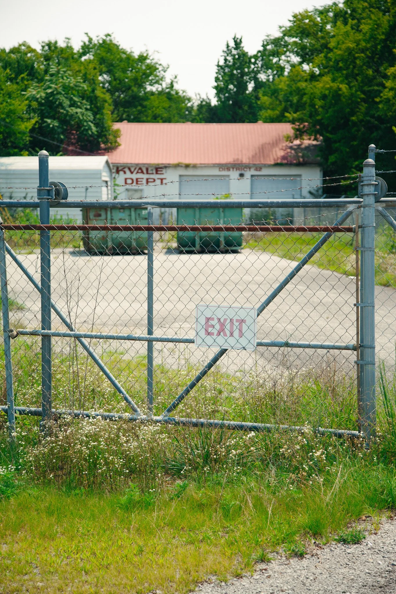 A chain link fence with a sign that says 'EXIT' in front of a gravel and grassy area, with a building labeled 'ROCKVALE FIRE DEPT' and 'DISTRICT 42' and trees in the background in Rockvale, TN