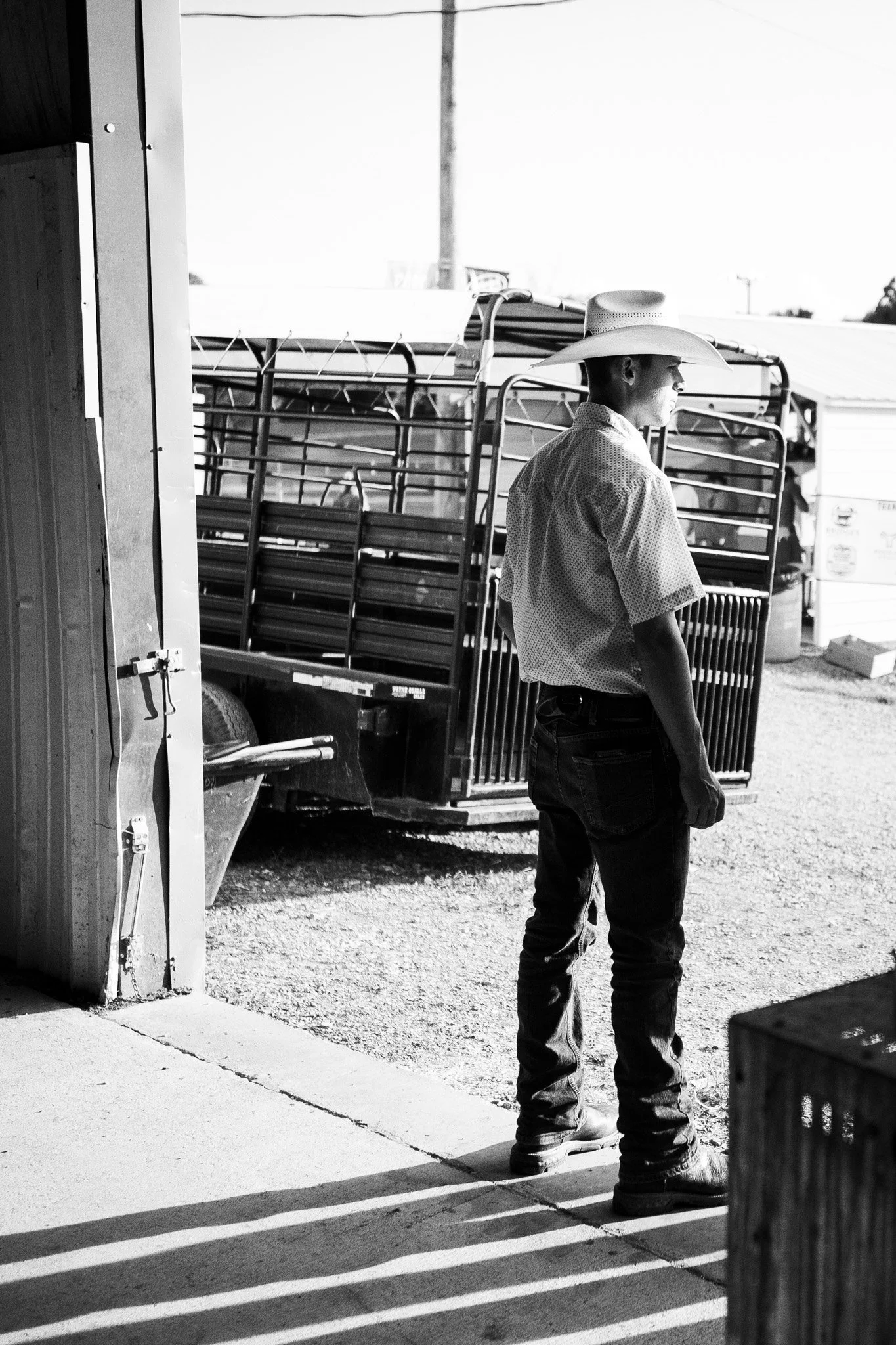 A man wearing a cowboy hat, a short-sleeved shirt, and jeans stands outside near a trailer with luggage racks. He appears to be looking into the distance, with sunlight casting shadows on the ground.