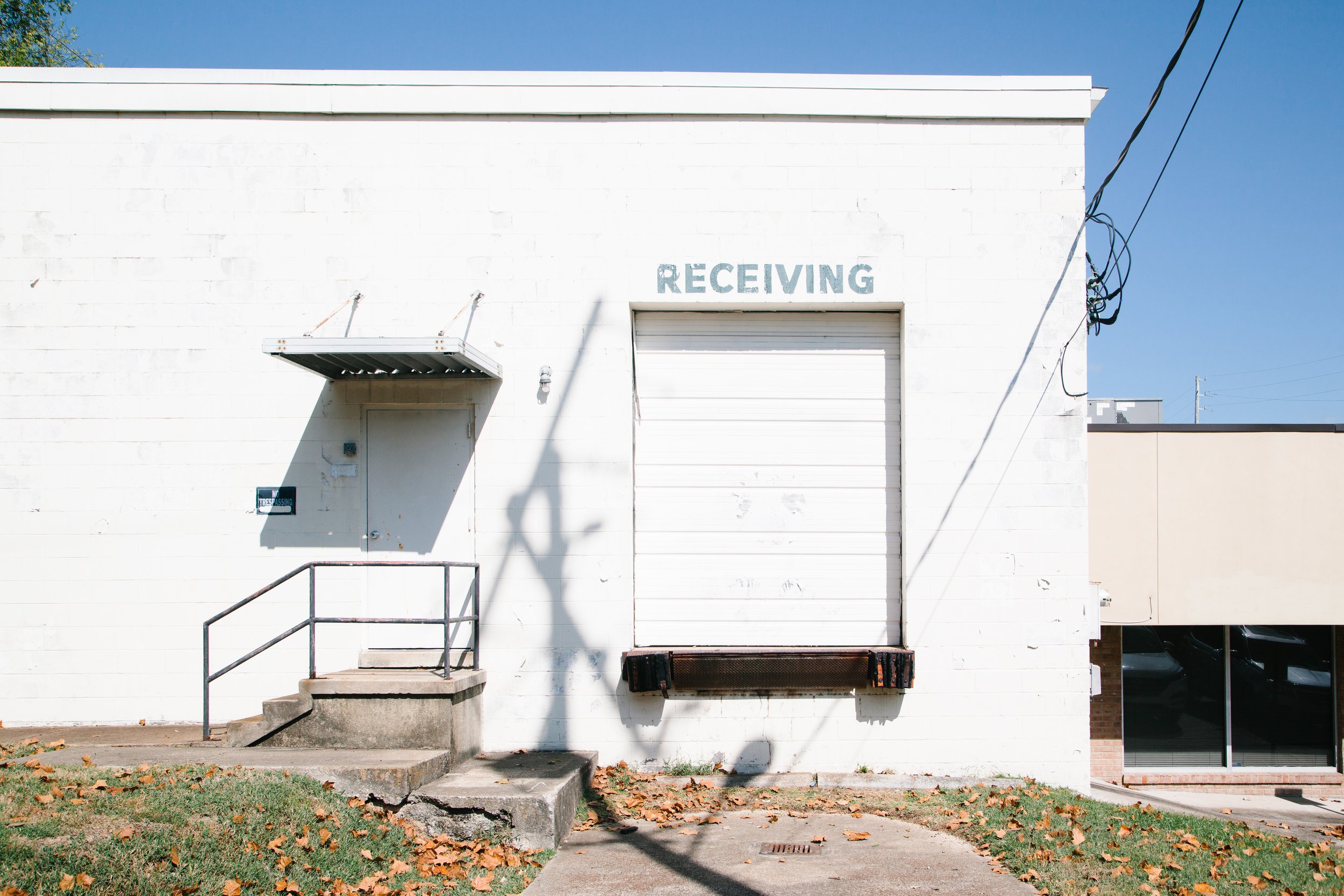 White building with a large roll-up door labeled 'Receiving,' a smaller door, a metal awning, a set of stairs, a handrail, a utility box, a sign that says 'No Trespassing,' a window, and electrical wires against a clear blue sky. Fallen leaves are sc
