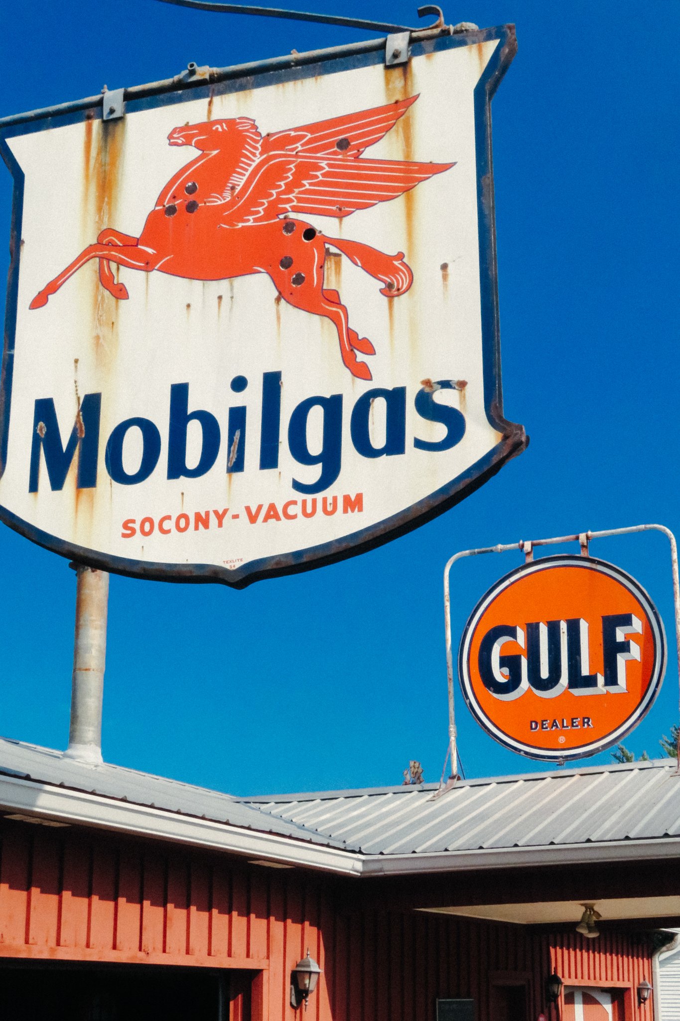 A vintage station wagon sign featuring a red Pegasus logo and the words 'Mobilgas' with 'Socony-Vacuum' underneath. The sign is rusty with a vertical streak of rust running down it. Adjacent is a circular Gulf logo sign on a beige building with a red