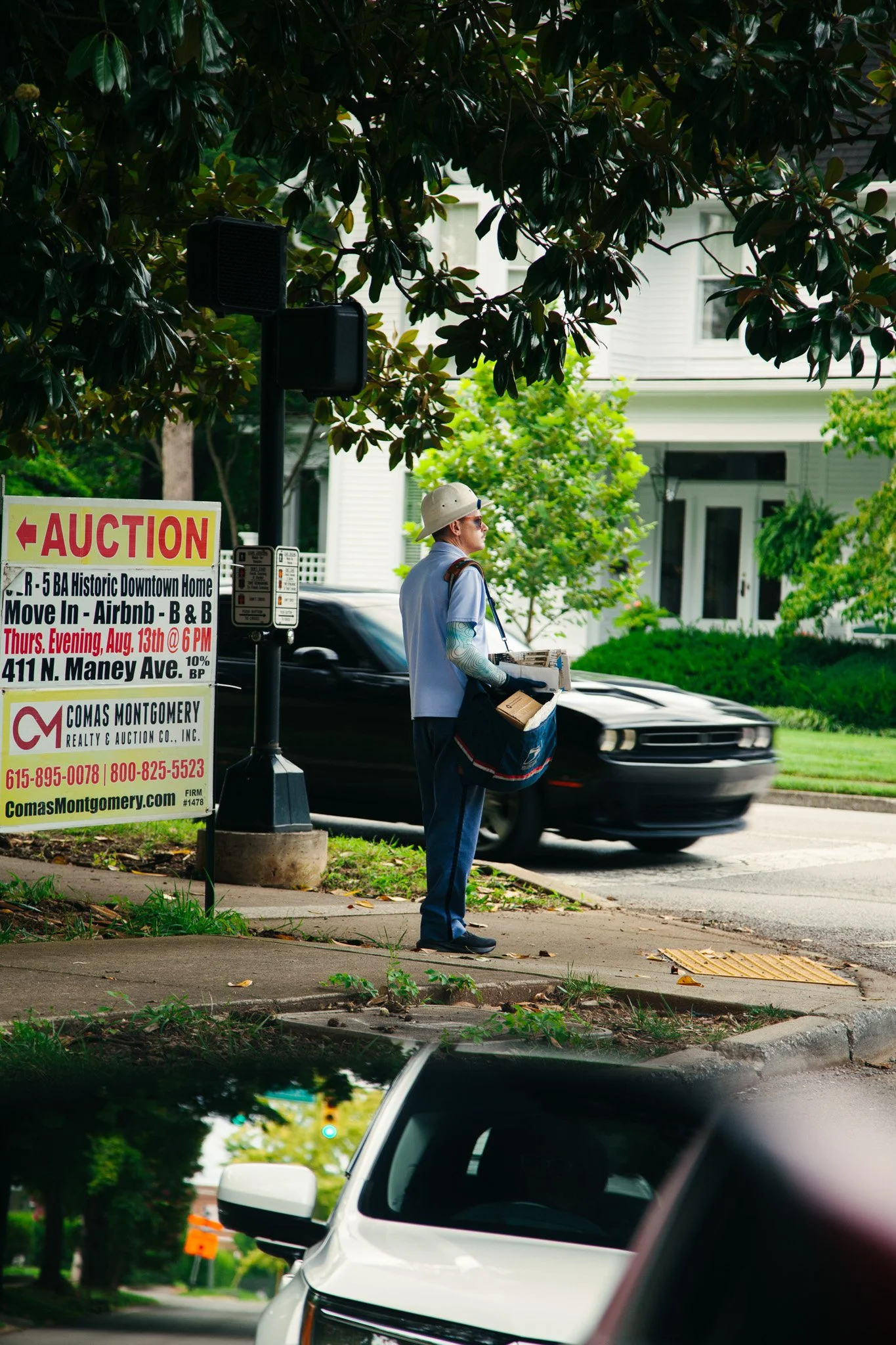 A person wearing a light-colored shirt, a wide-brimmed hat, and sunglasses stands on a sidewalk, holding a brown paper bag and some boxes. A black car passes by on the street. There are trees, a white house with windows, and a sign advertising an auc