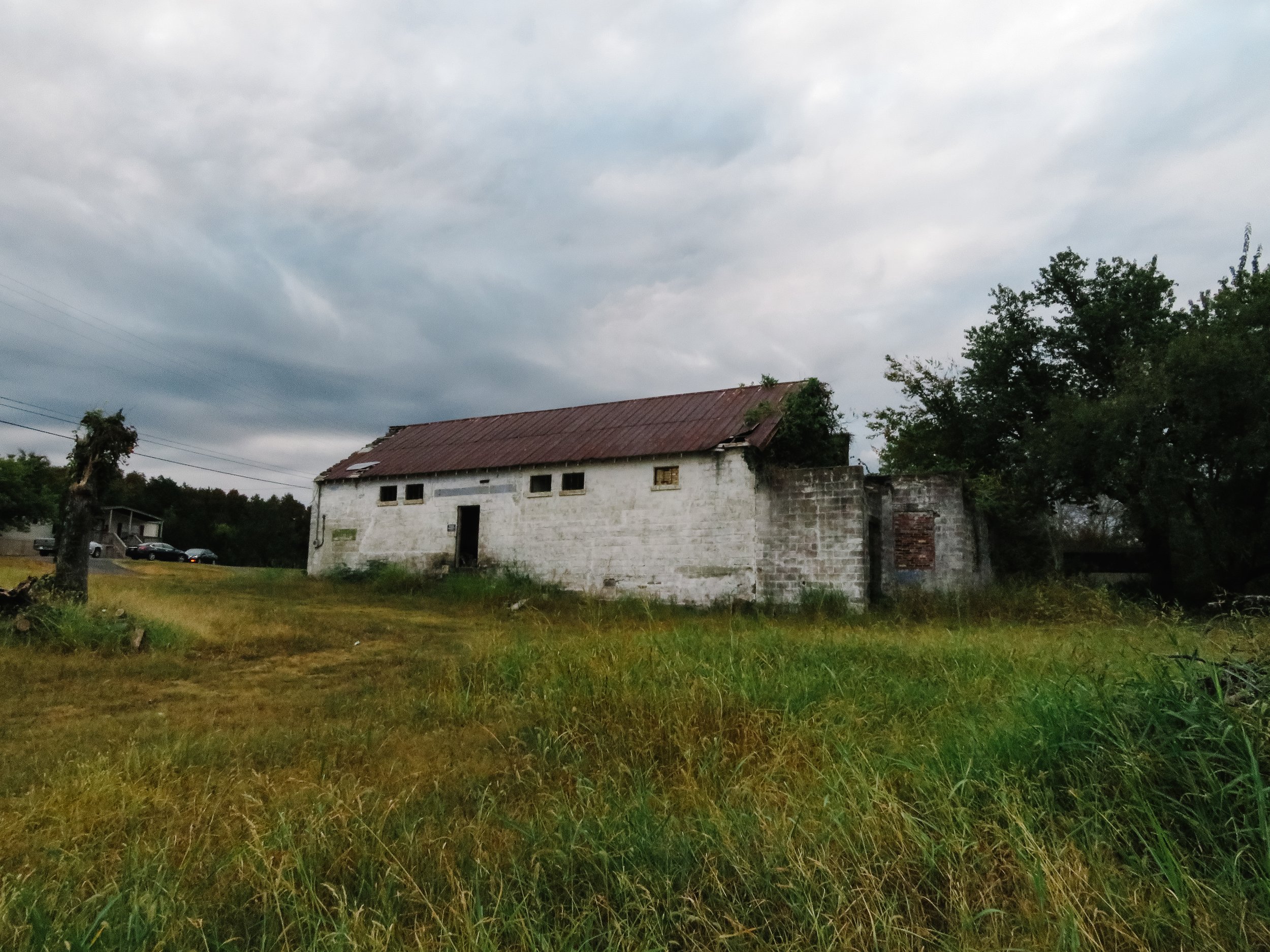 An old, weathered white building with a rusted metal roof and multiple small windows. The building is surrounded by tall grass and trees, with a cloudy sky overhead.