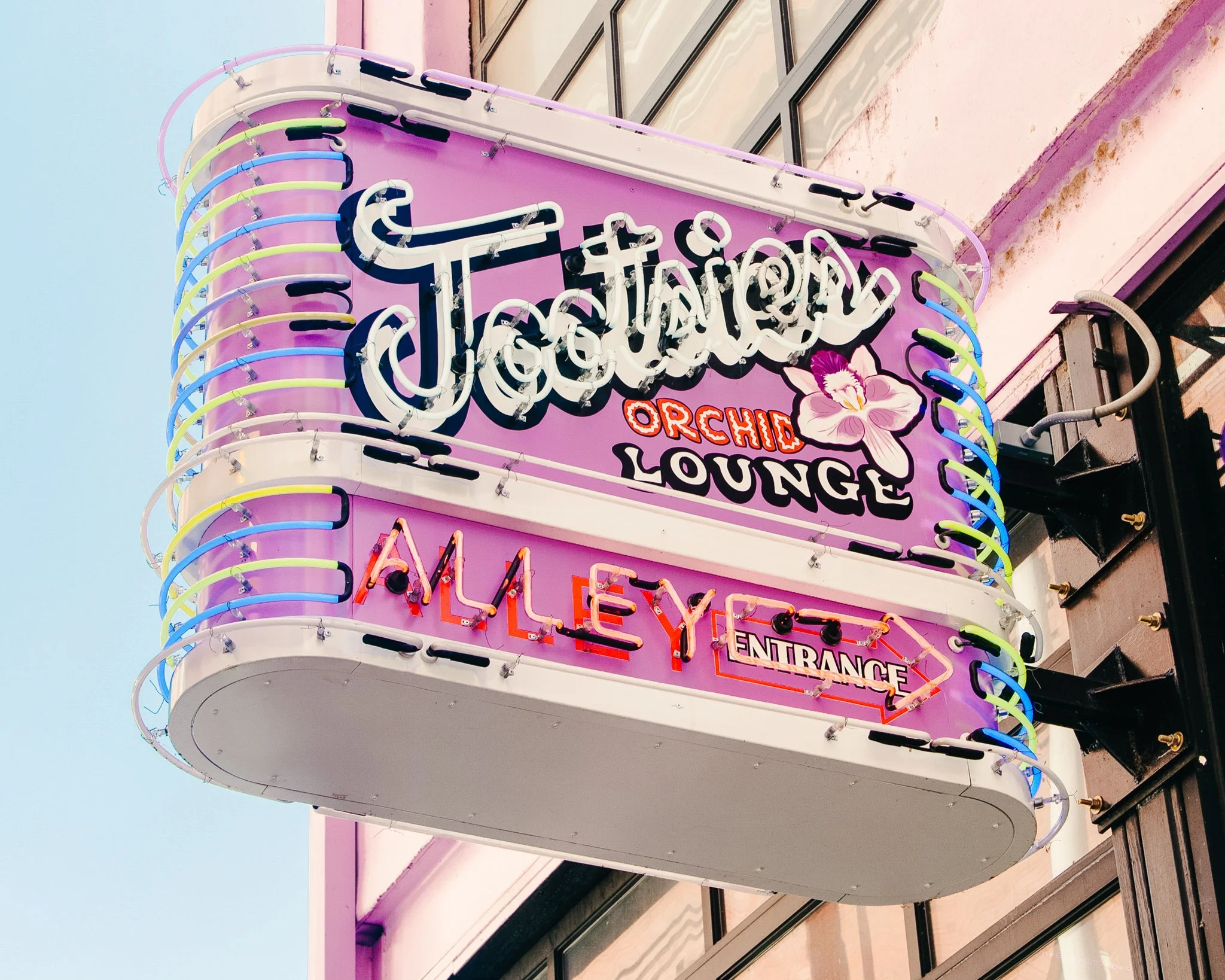 Neon sign for a lounge with pink, blue, green, and white colors, featuring a flower illustration and text in various styles, attached to a pink building facade.