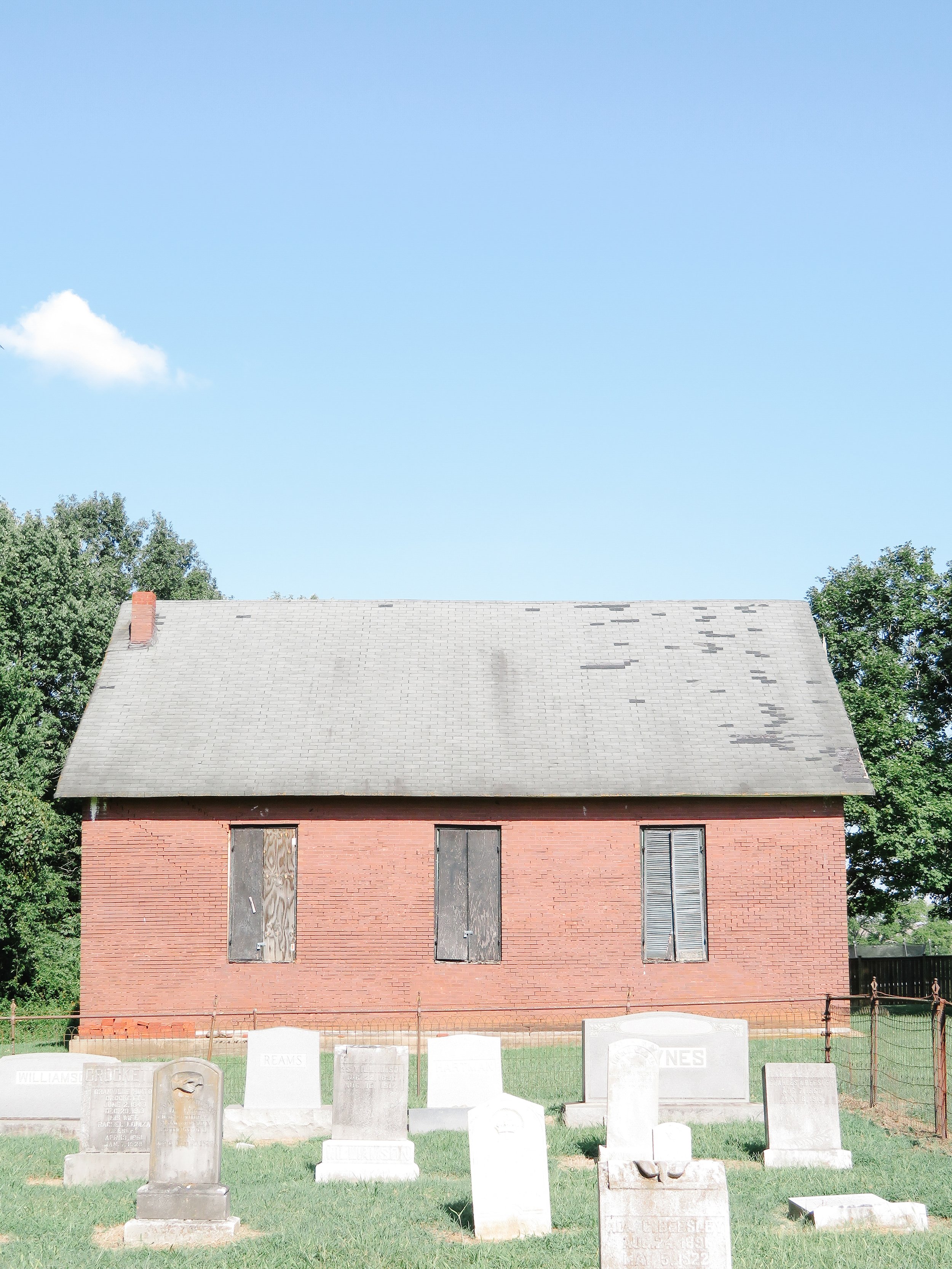 A brick building with a sloped gray roof and boarded-up windows, surrounded by a grassy area with several headstones in front, some marked with names like Williams and Crews, and trees on either side. The sky above is clear and blue with a few clouds