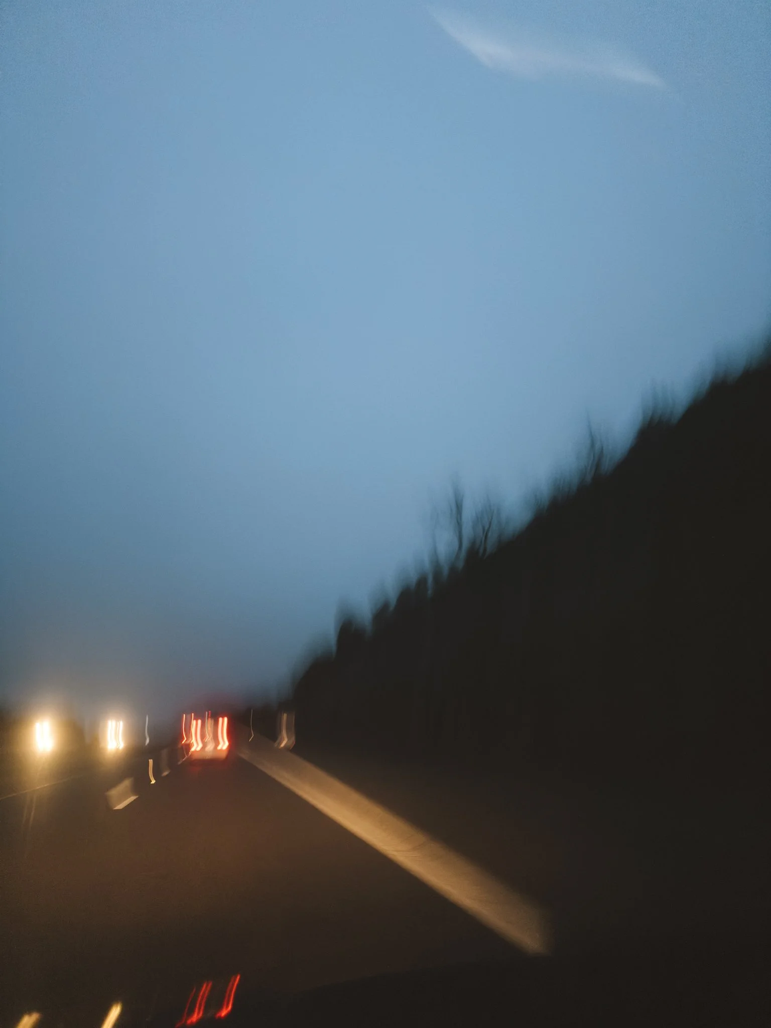 Nighttime blurred image of a highway with vehicle lights, distant city lights, and dark hills in the background.