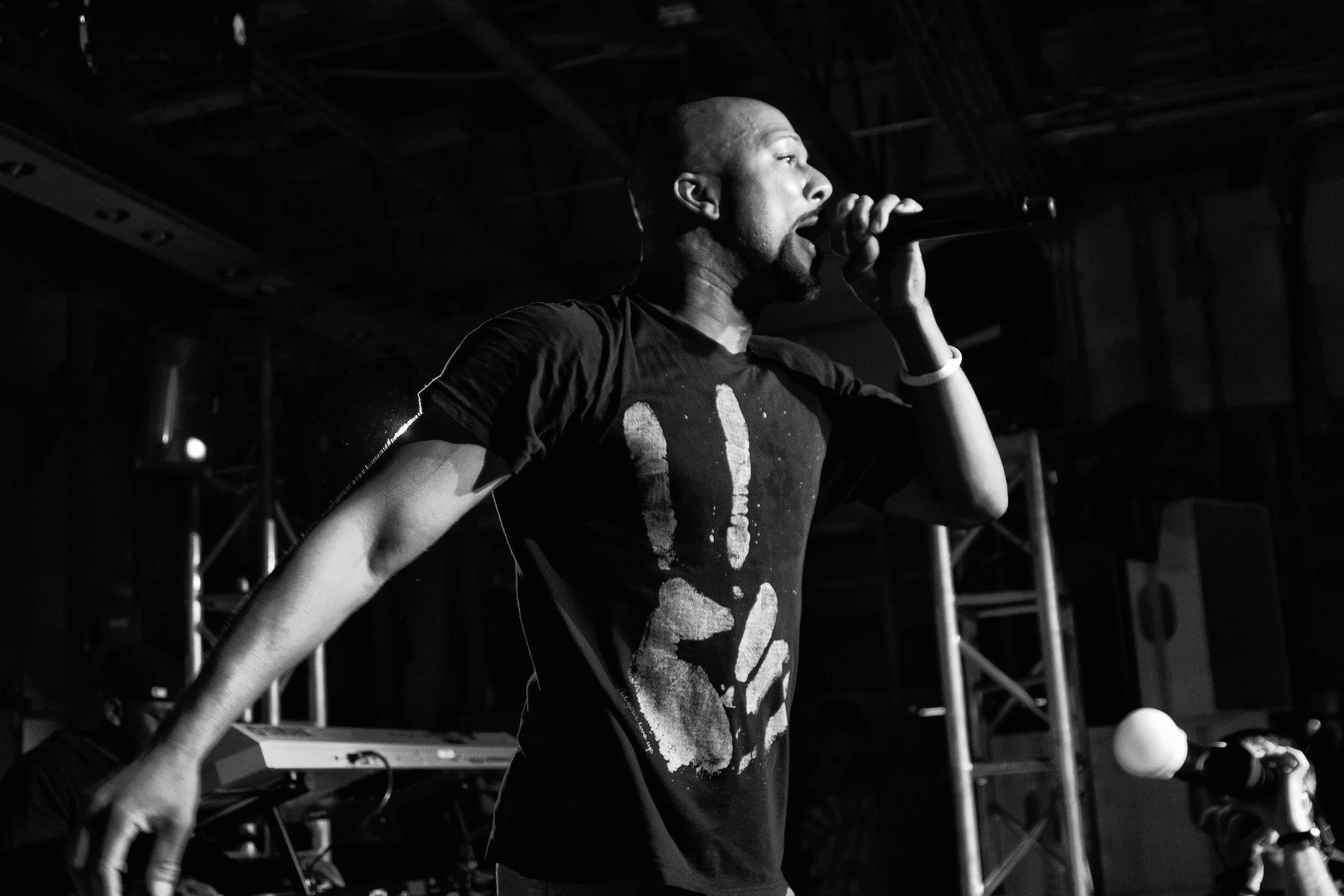 A black-and-white photo of a male performer singing into a microphone on stage. He is wearing a dark t-shirt and appears to be passionately engaged in his performance, with a focused expression. The background includes stage equipment and lighting st