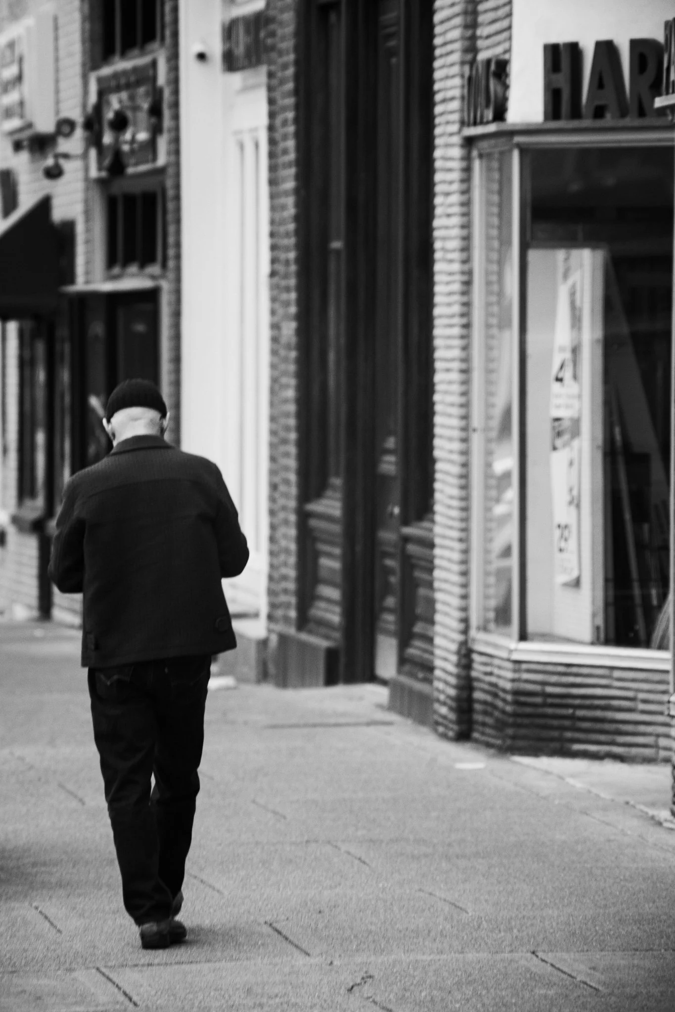 A person with a black beanie and dark clothing walking on a city sidewalk, with storefronts and large windows visible on the right side of the street. The photo is in black and white.