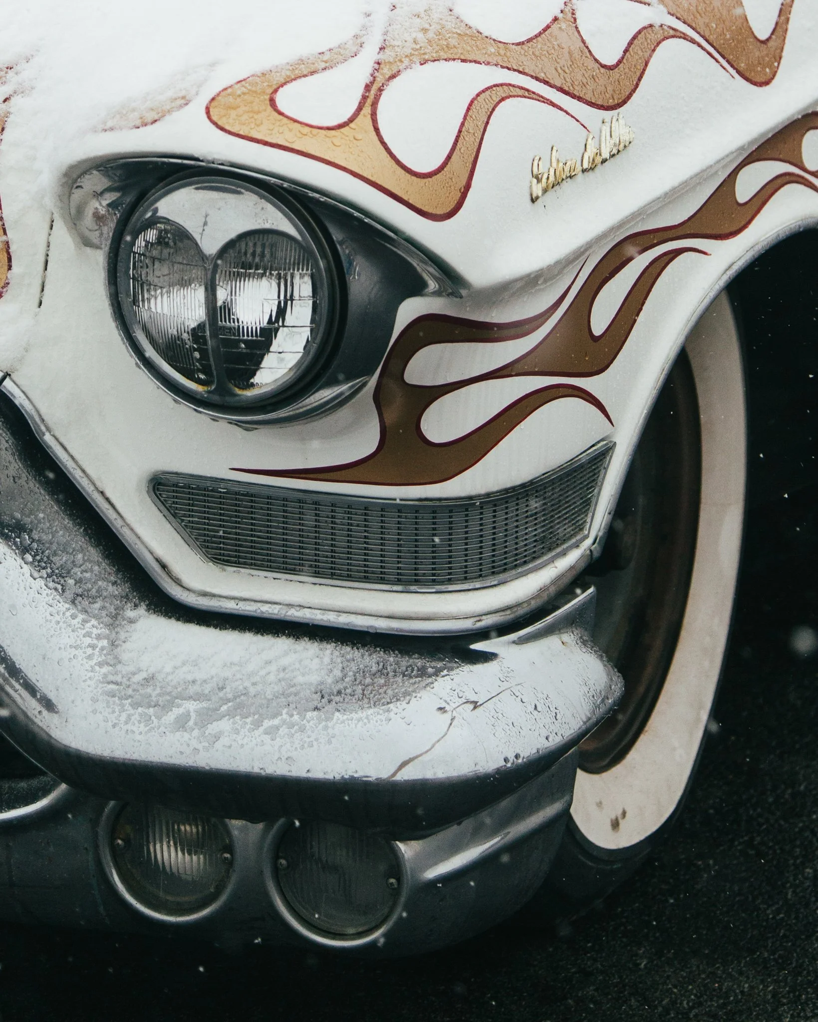 Close-up of a vintage white car with flames painted in brown and red, showing the front left side including the headlight, grille, and part of the hood with snow on it.