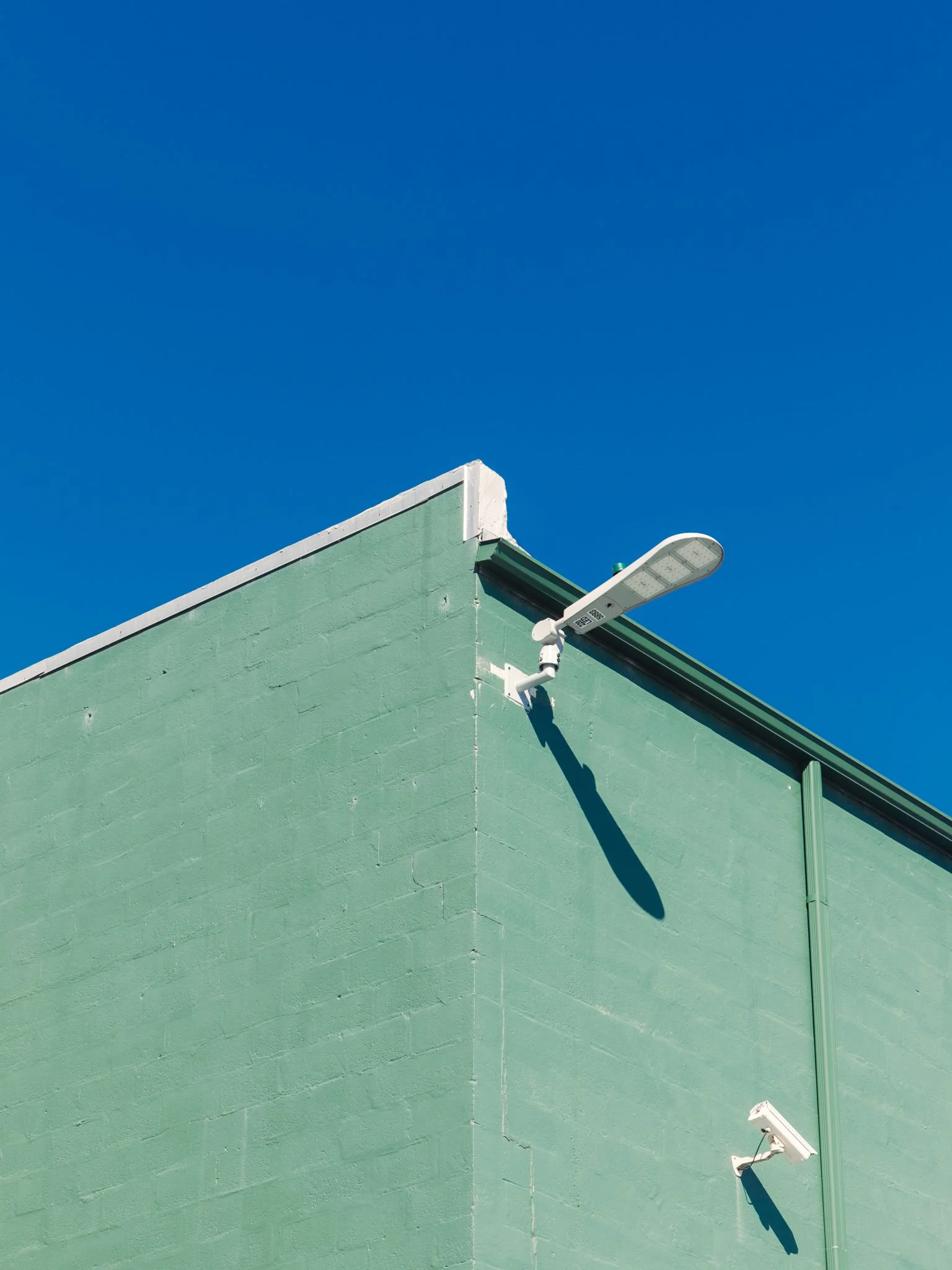 A green building corner with a light fixture casting a shadow on the wall against a blue sky.