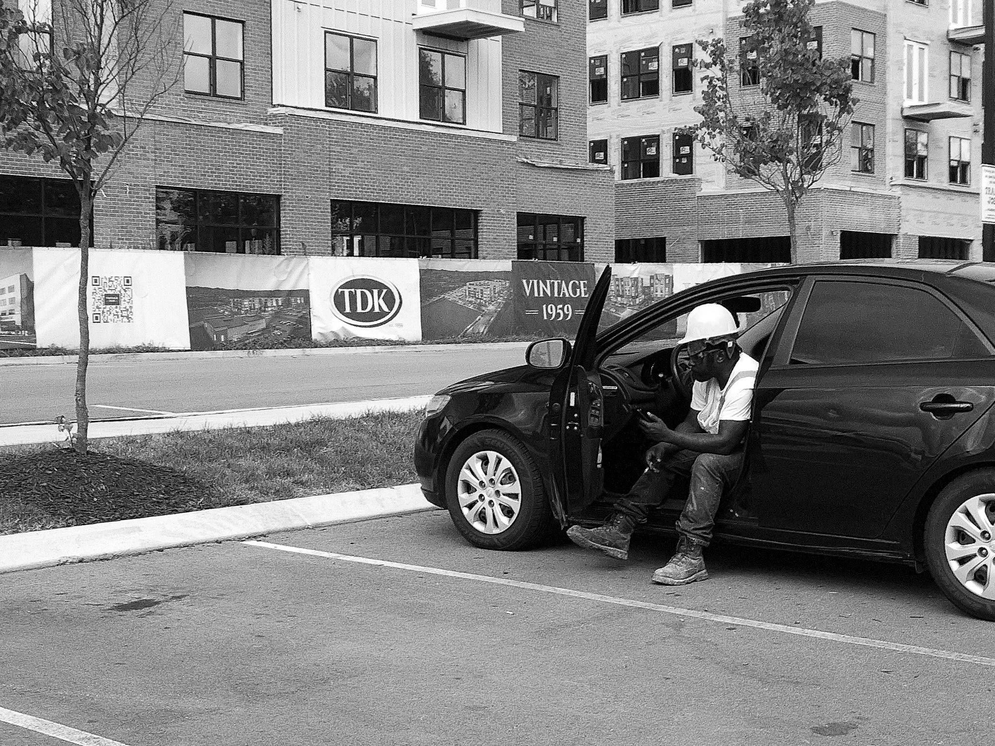 A man is sitting inside a parked black car with the door open, wearing a helmet and looking at a smartphone.