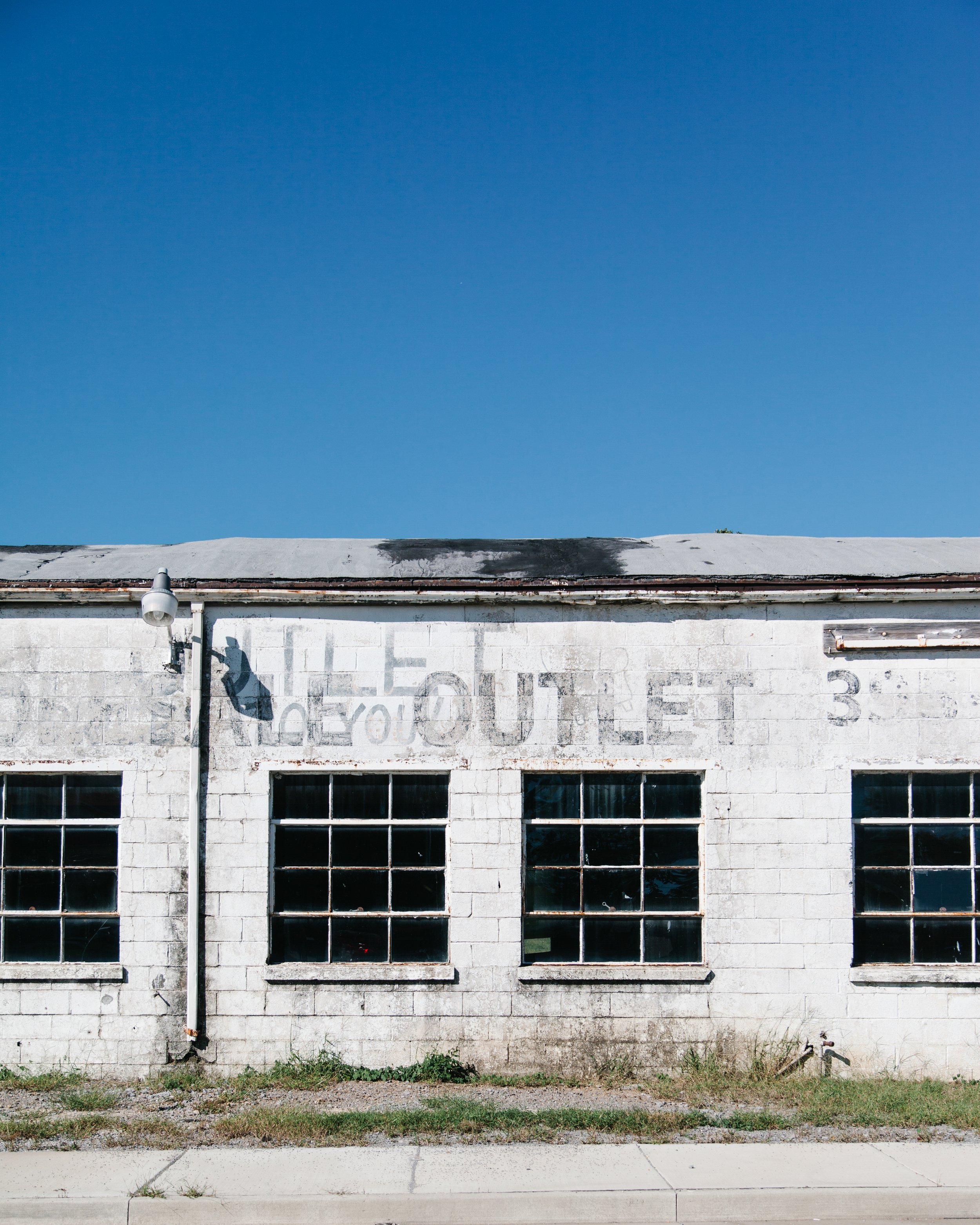 An old abandoned commercial building with large grid windows and a faded, partially visible sign painted on the wall. The building has a flat roof with some black streaks and a small patch of grass and sidewalk in front.