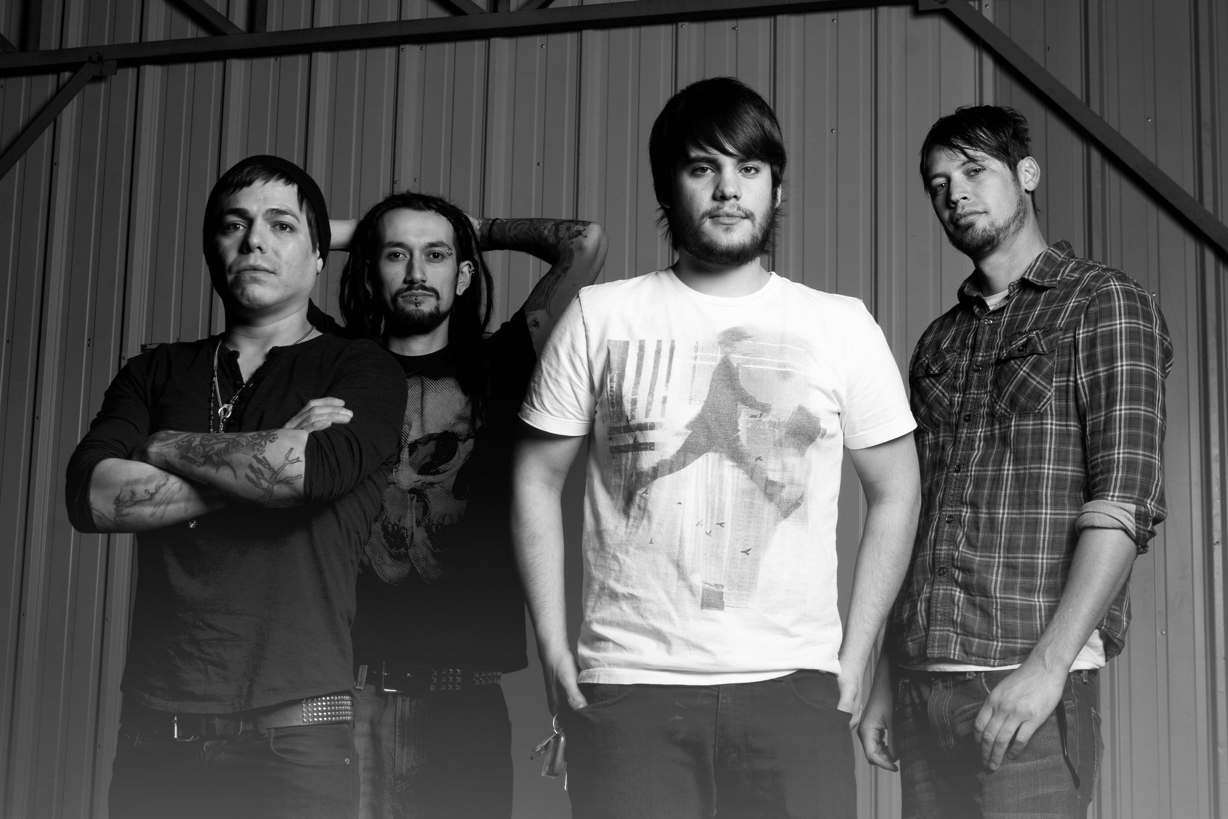 A black-and-white photo of four men standing in front of a metal wall, with serious expressions, some with crossed arms or relaxed postures.
