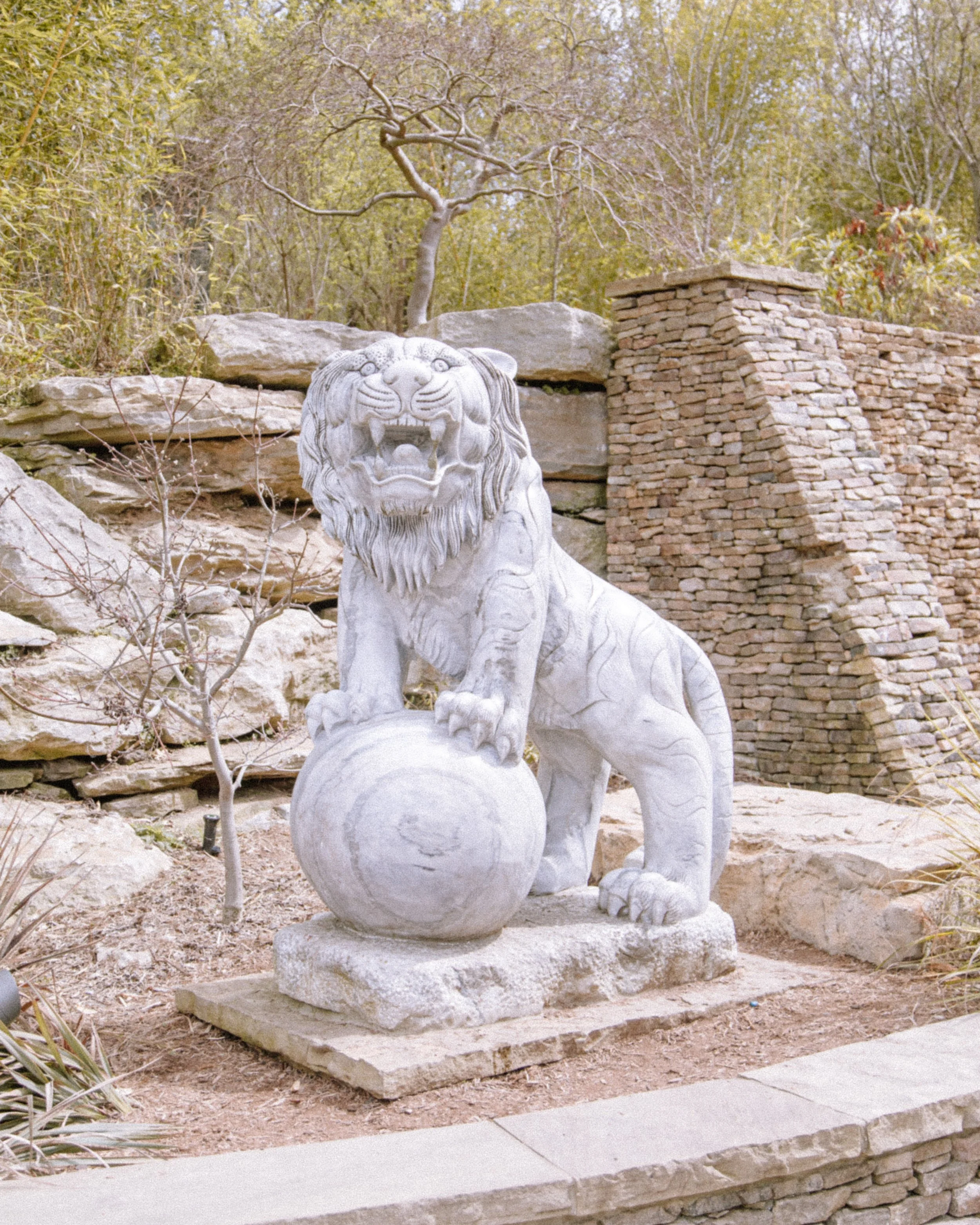 Stone lion statue with an orb in front, set on a concrete base, with layered stone wall and trees in the background.