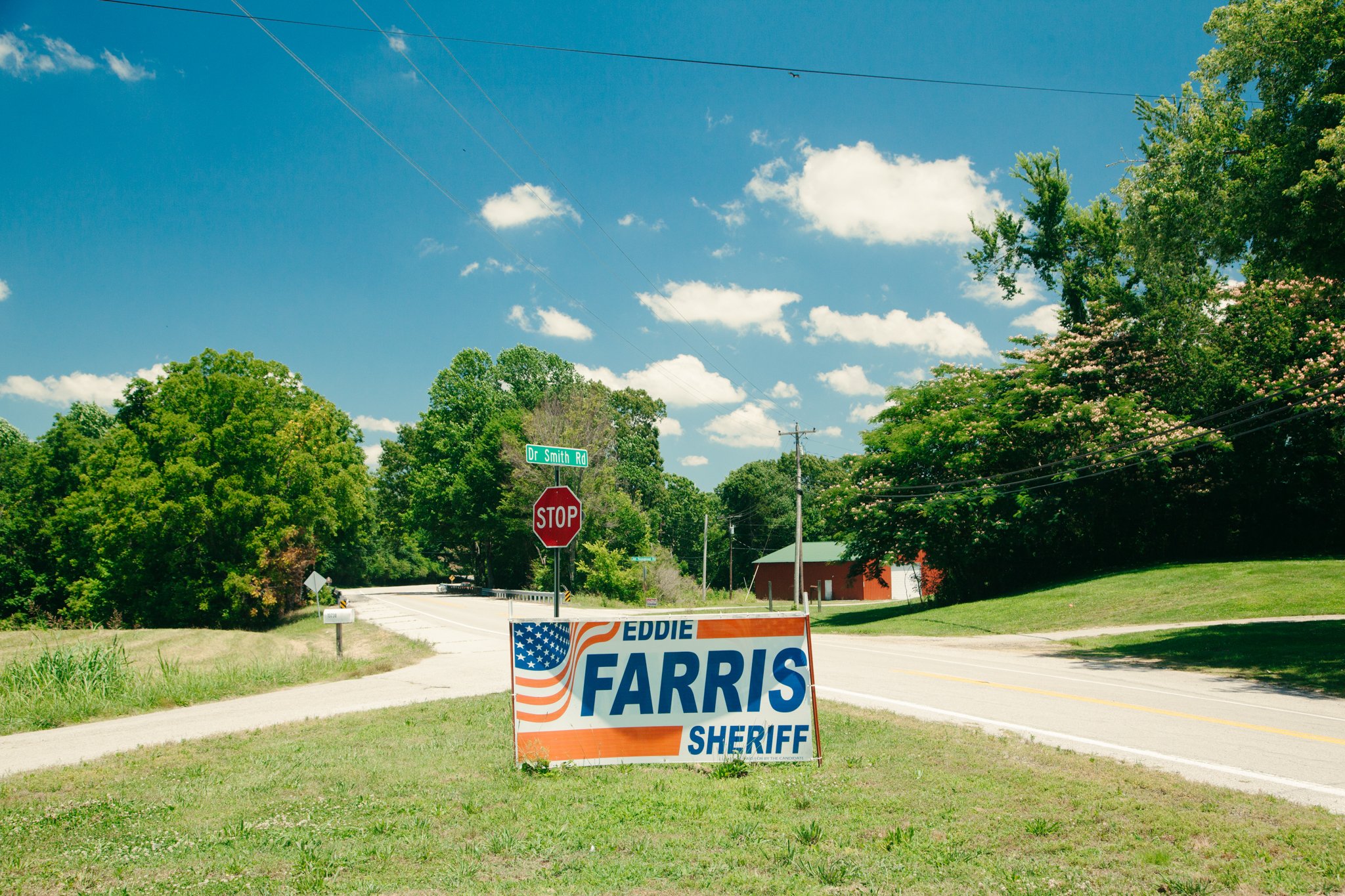 A rural street scene with a campaign sign for Eddie Farris for sheriff. There is a stop sign, a street sign for Dr. Smith Rd, trees, a sidewalk, and a blue sky with some clouds.