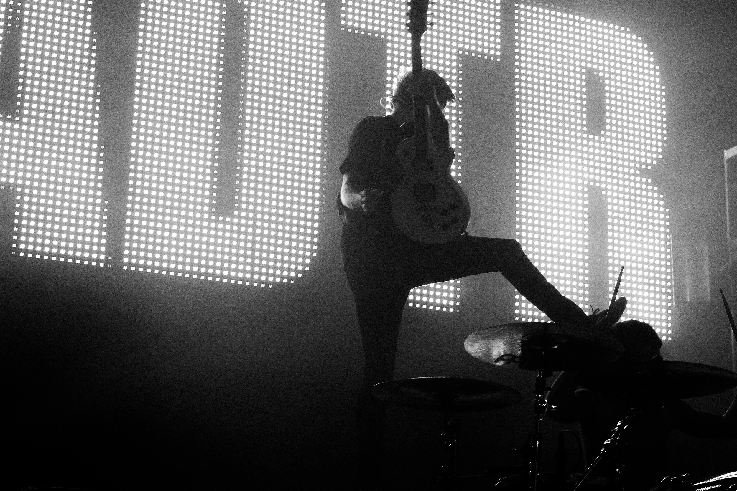 A musician playing electric guitar on stage with large illuminated LED panels in the background, black and white photo.