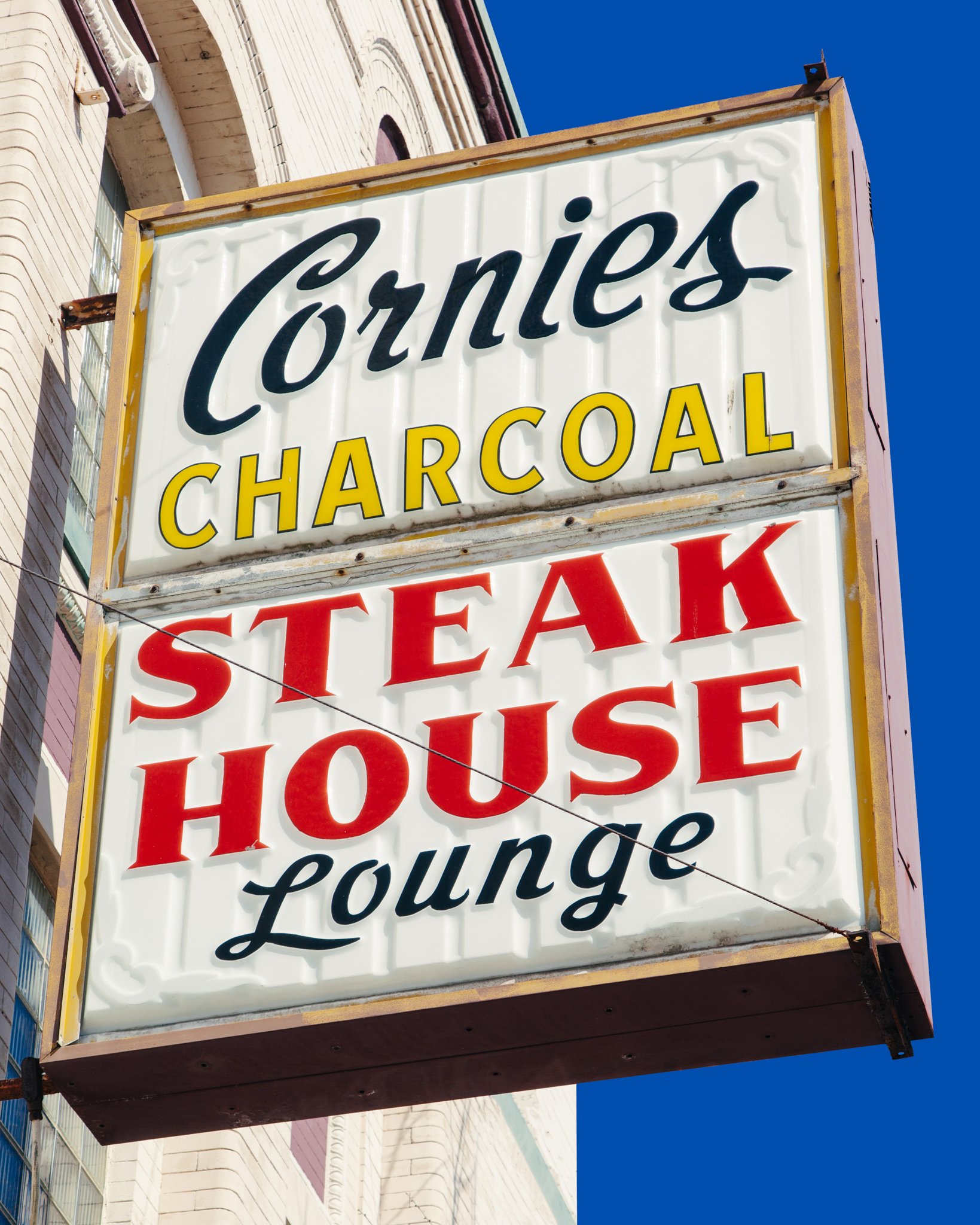 A vintage theater marquee with a white background displays the words 'Cornies Charcoal' in black cursive and yellow block letters, respectively. The marquee is mounted on a building with beige brick architecture and is set against a clear blue sky.