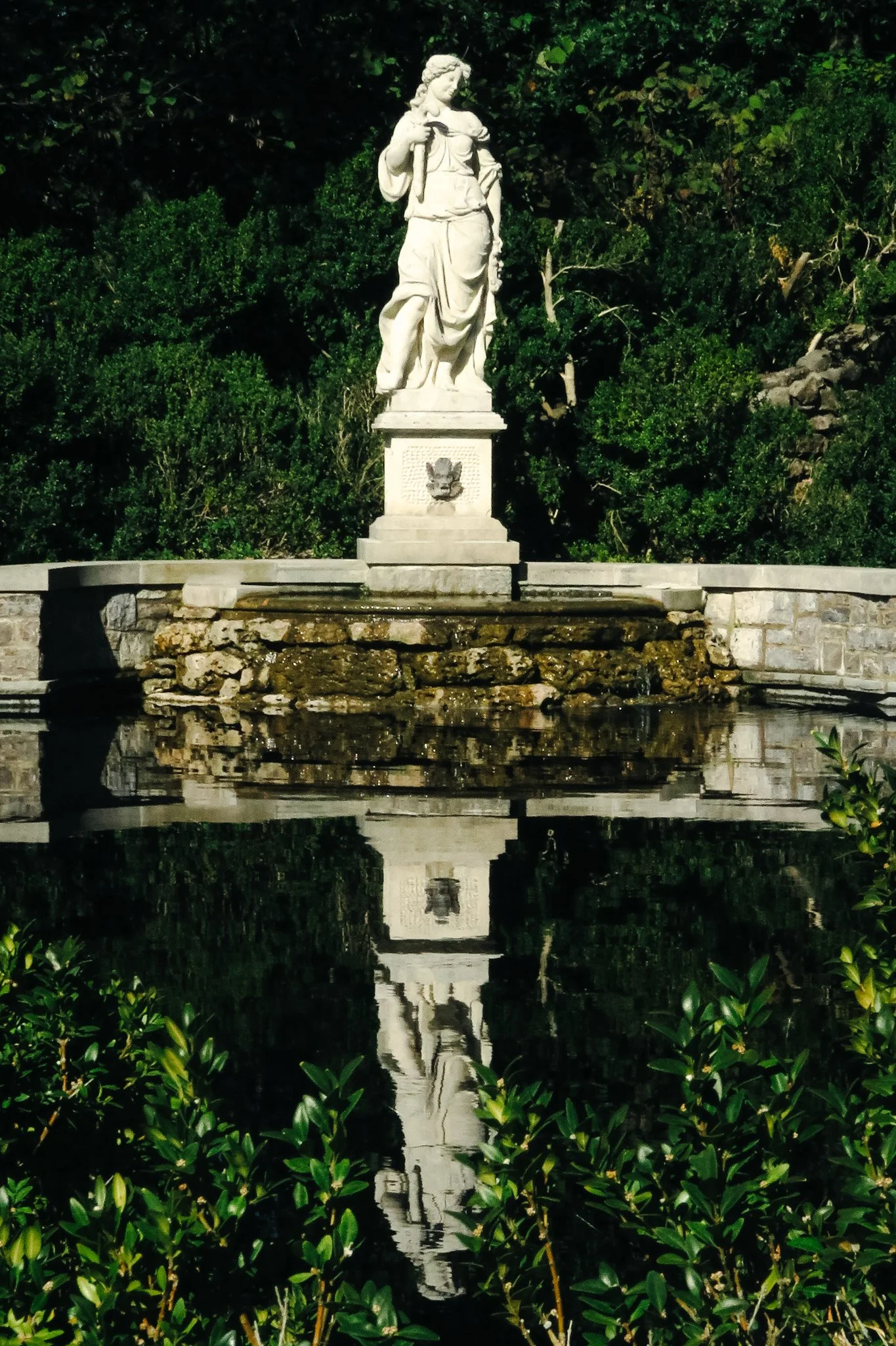 A white stone statue of a woman holding a sword in one hand, standing on a pedestal, with lush green bushes and a fountain with water and a reflection in the foreground.
