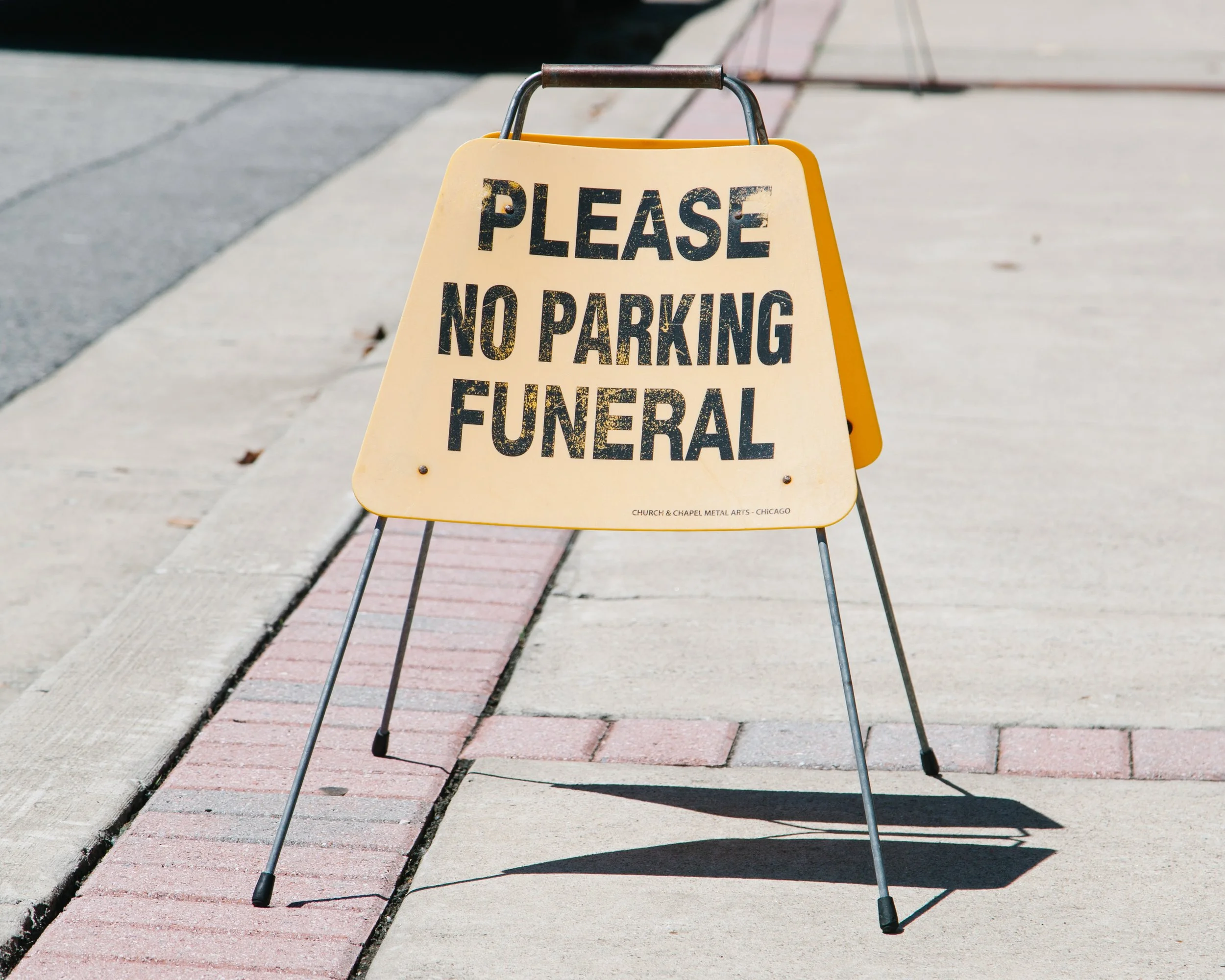 A yellow protest sign on a sidewalk that reads 'Please No Parking Funeral' with a small note at the bottom indicating it is by Church & Chapel Metal Arts in Chicago, placed on a brick-lined sidewalk.