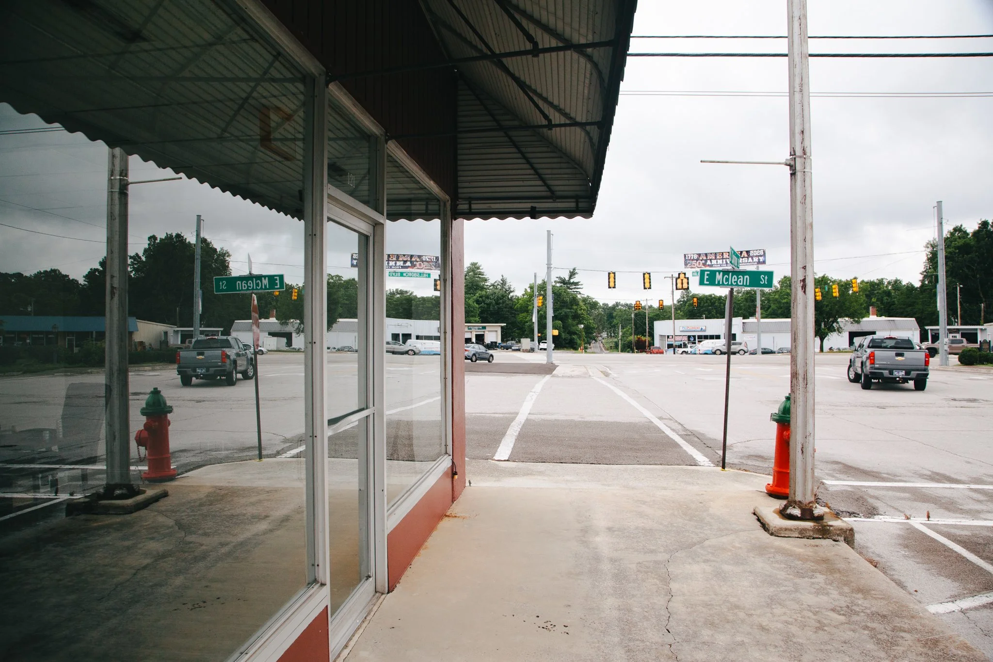 A view of a parking lot from inside a small storefront or restaurant with a glass front door. The street signs indicate the corner of E McLean Street and E Melton Street. Several cars are parked in the lot, and businesses and trees are visible in the