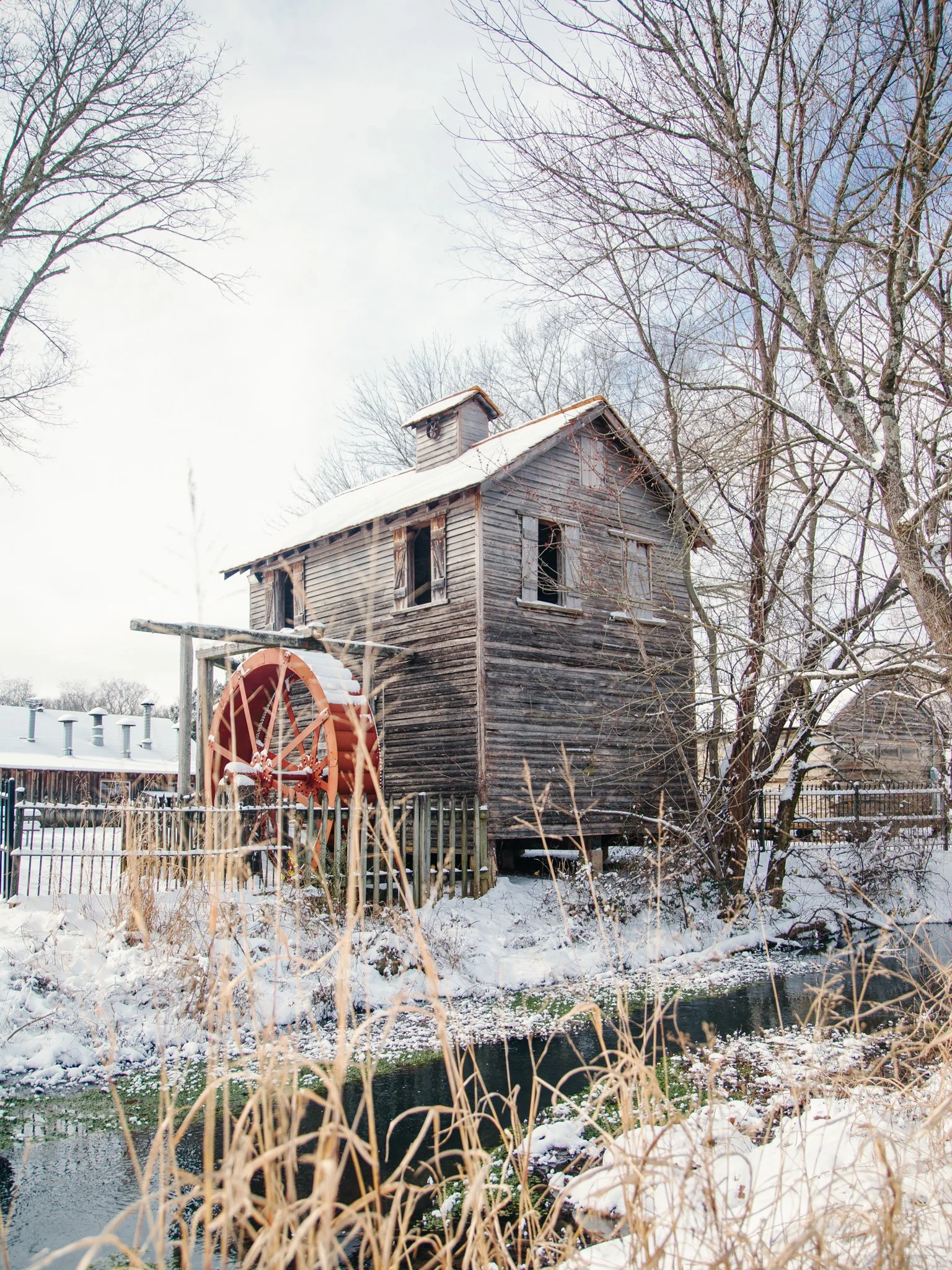 A weathered wooden granary with a small loft and a water wheel in a snowy landscape, surrounded by leafless trees and a small creek.